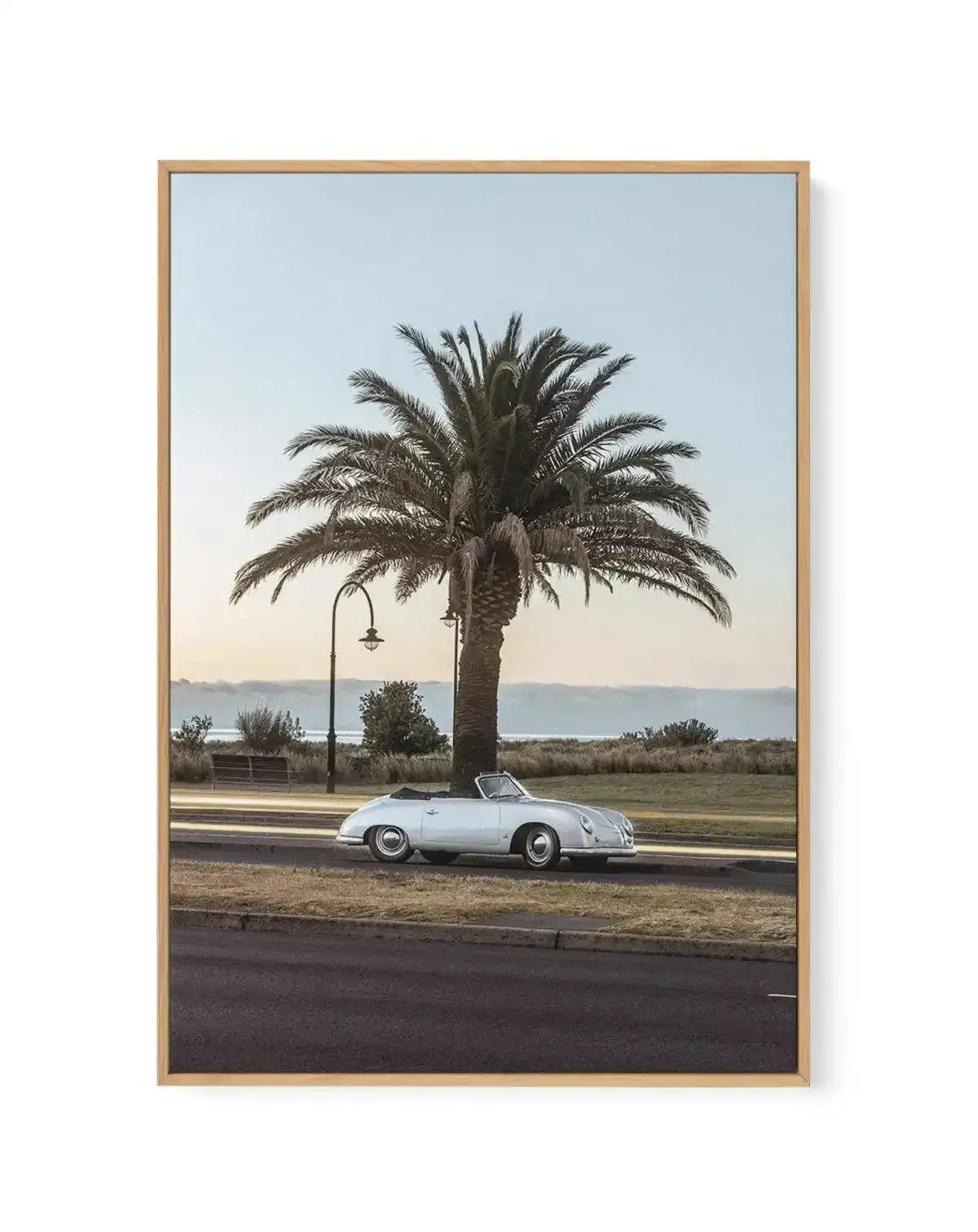 Framed canvas art print featuring a vintage white convertible sports car parked beneath a large palm tree on a coastal promenade. A street lamp stands nearby, with a wooden bench visible in the background. The scene overlooks a calm sea under a soft, hazy sky in muted blue and cream tones. The photograph has a nostalgic, mid-century aesthetic with natural lighting. The canvas is mounted in a natural wood frame with a white border.