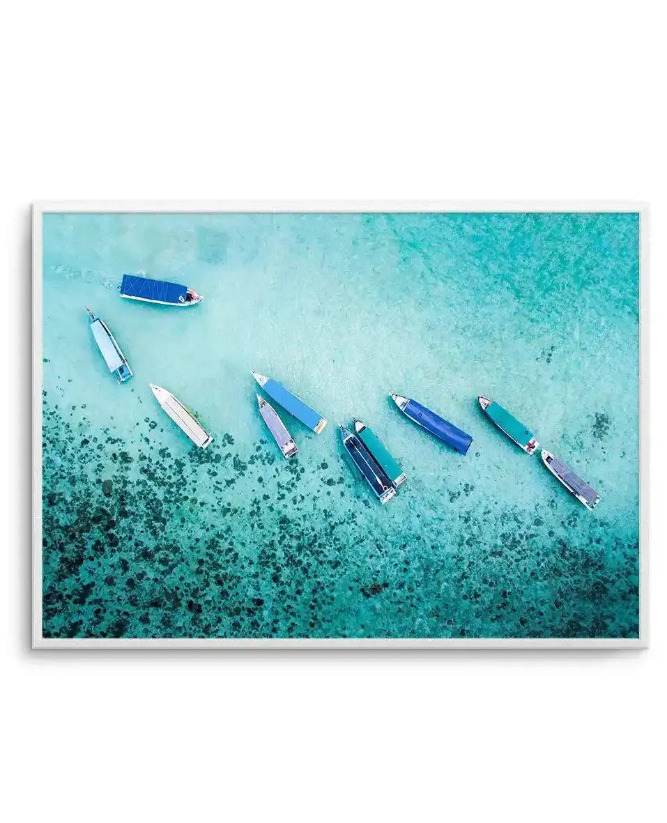 Aerial photograph art print showing eight fishing boats scattered across shallow turquoise water with darker teal seagrass visible below. Boats feature blue, white, and dark hulls with small red accents. Upper left shows deeper turquoise water, lower left transitions to darker seafloor vegetation. Print is framed in white with white border, horizontal landscape orientation.