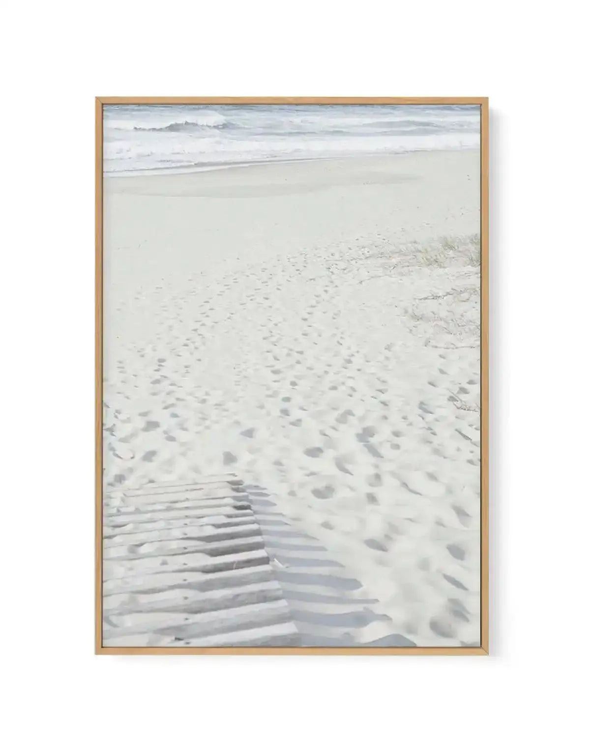 Framed canvas art print with natural wood frame featuring a serene beach scene. The composition shows footprints and tire tracks in pale sand in the foreground, with gentle waves and white foam in the middle distance, and a misty gray-blue ocean with distant hills under an overcast sky in the background. The photograph has a soft, muted color palette of cream, pale gray, and cool blue tones, creating a calm, contemplative coastal mood.