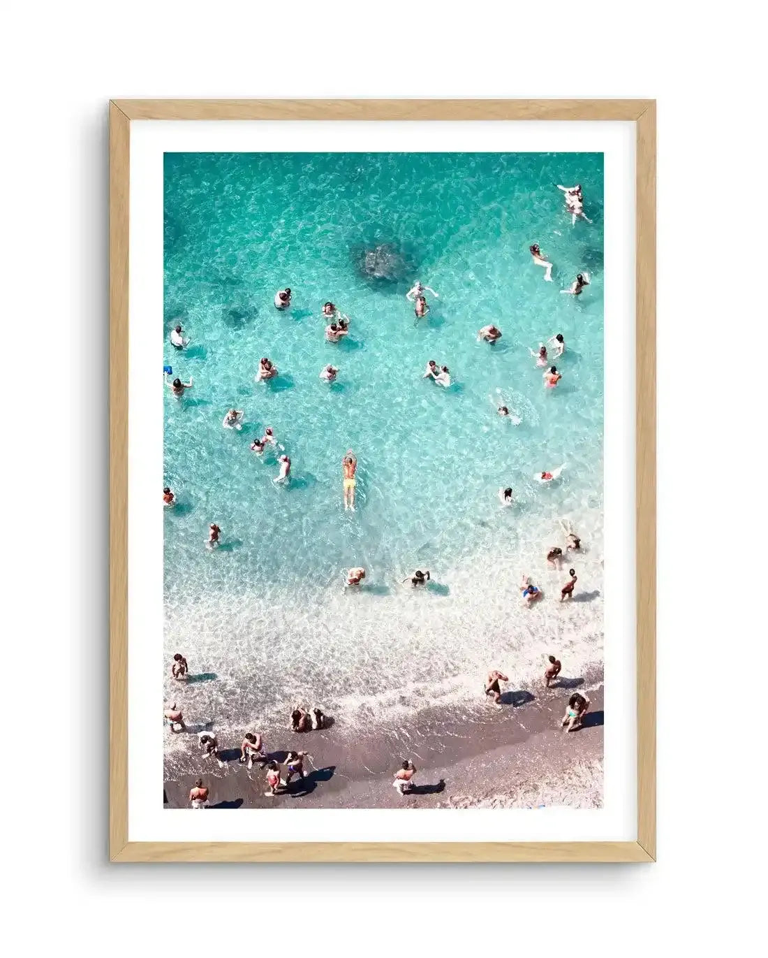 Framed aerial photograph of a vibrant beach scene with a natural wood frame and white border. The image captures numerous people swimming in clear turquoise water and relaxing on the light brown sandy shore, with gentle waves lapping at the edge.