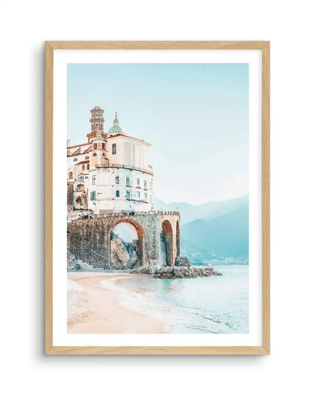 Framed photography art print of a coastal town with a natural wood frame and white border. The composition features a white building with a green dome and bell tower perched on a stone wall with arched openings, overlooking a sandy beach and calm turquoise sea. Distant mountains are visible under a pale blue sky.