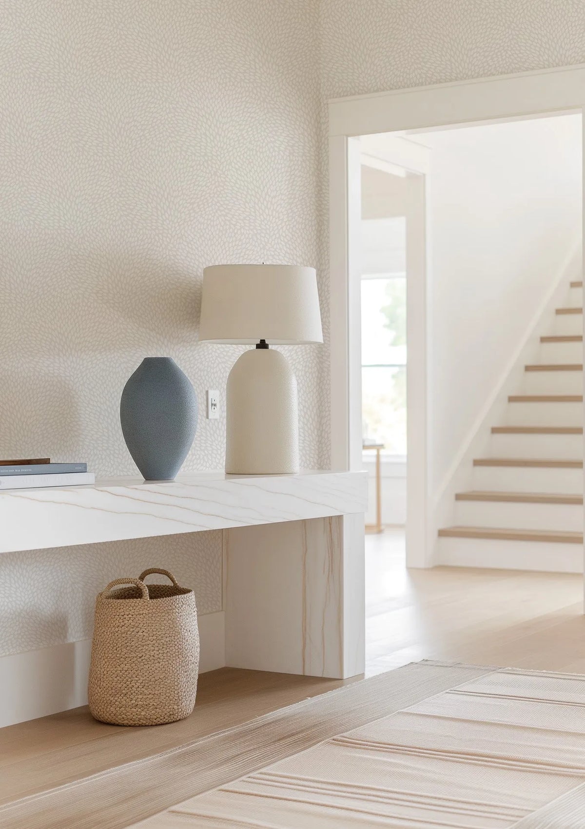 Lifestyle shot featuring a hallway with light beige wallpaper patterned with small, organic, off-white leaf-like shapes. A white console table with subtle grey veining holds a tall, textured grey vase, a stack of books, and a cream-colored table lamp. A woven basket sits on the floor beneath the console. In the background, a doorway opens to a bright space with a wooden staircase.