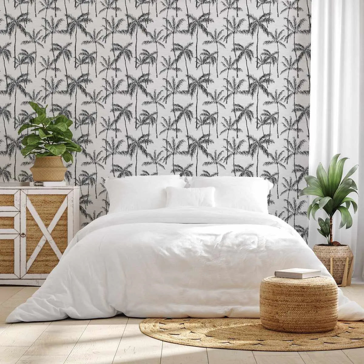 "Bedroom with a tropical-themed wallpaper featuring a repeating pattern of black palm trees on a white background. The room includes a bed with white linen bedding, a light wood and woven rattan bedside cabinet with a green plant in a woven basket, a round woven jute rug, and a woven jute ottoman with a book on top. A tall green plant in a woven basket stands next to white curtains on the right side of the bed."