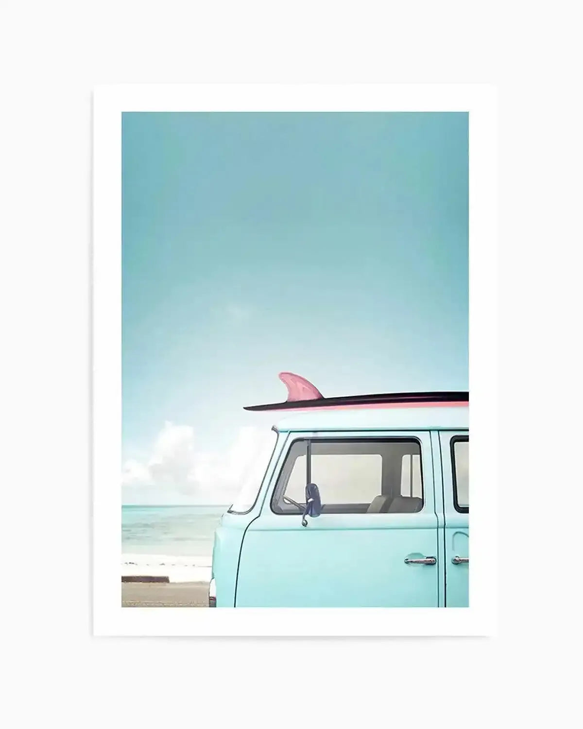 Framed photography art print featuring the front of a light blue vintage Volkswagen Kombi van with a pink surfboard on its roof, parked on a sandy beach next to the turquoise ocean under a clear blue sky. The print is displayed in a white frame with a white border.