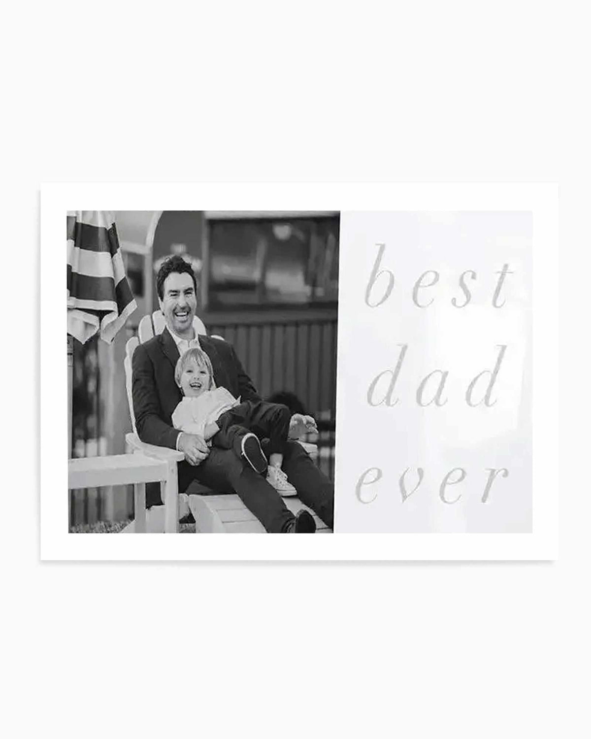 Black and white art print featuring a custom photo design. Left side displays a black and white photograph of a smiling man in a dark blazer holding a young child, seated on white porch steps with an American flag visible in soft focus behind them. Right side features the text "best dad ever" in a light gray, elegant script font. The print has a clean, minimalist layout on a white background, creating an emotional Father's Day or family keepsake design.