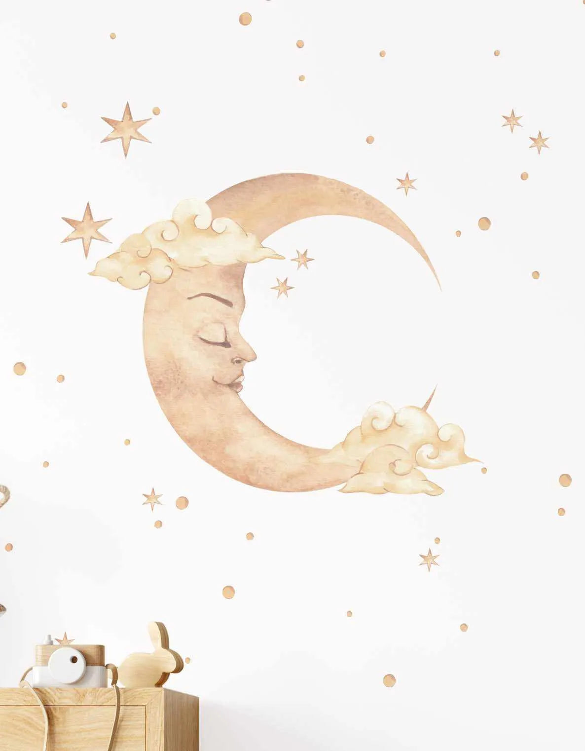 Wall decal set featuring a large crescent moon with a sleeping face, rendered in a soft watercolour style with warm beige and light brown tones. The moon is surrounded by whimsical clouds in similar hues and scattered small stars and dots in a golden-beige colour, all against a plain white background. A wooden toy camera and bunny figurine are visible in the bottom left corner, resting on a light wood dresser.