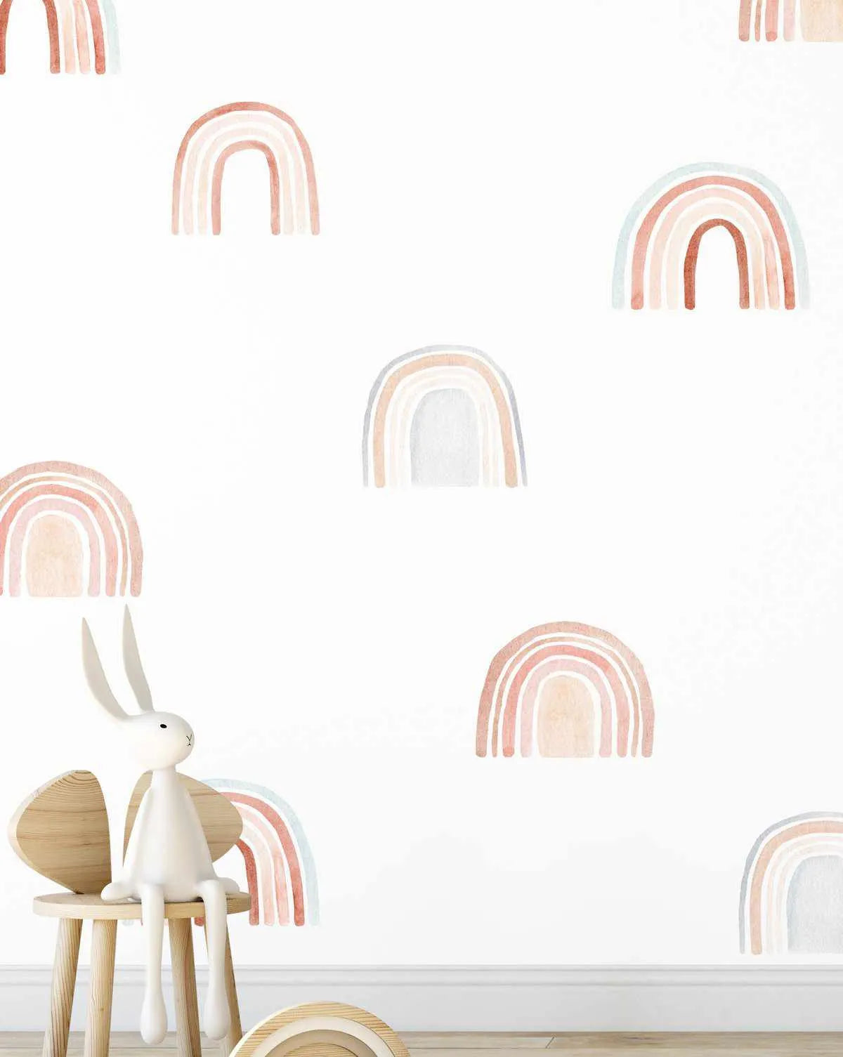 Peel and stick wall decal set featuring multiple hand-painted watercolour rainbows in muted pastel shades of dusty rose, pale peach, soft grey, and light blue on a white background. The rainbows vary in size and are scattered across the wall, with a white rabbit toy sitting on a wooden stool in the foreground.
