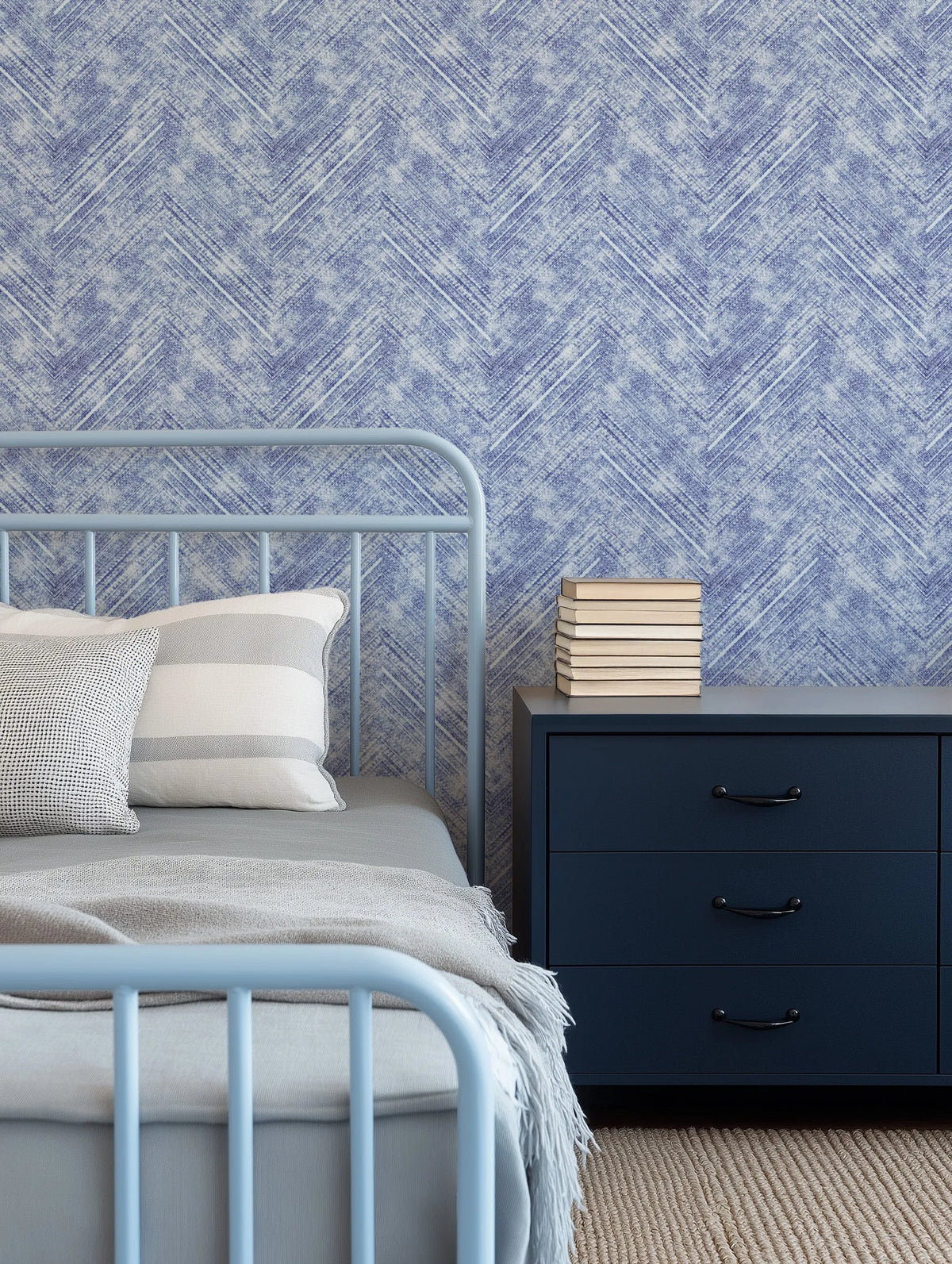 Lifestyle shot of a bedroom featuring a light blue metal bed frame with a grey duvet and throw, and two pillows (one white with grey stripes, one white with a black grid pattern). To the right, a dark blue three-drawer nightstand with black handles holds a stack of books. The wall behind the bed is covered in a denim blue and white chevron pattern wallpaper with a textured, distressed look. The floor is covered with a light brown woven rug.