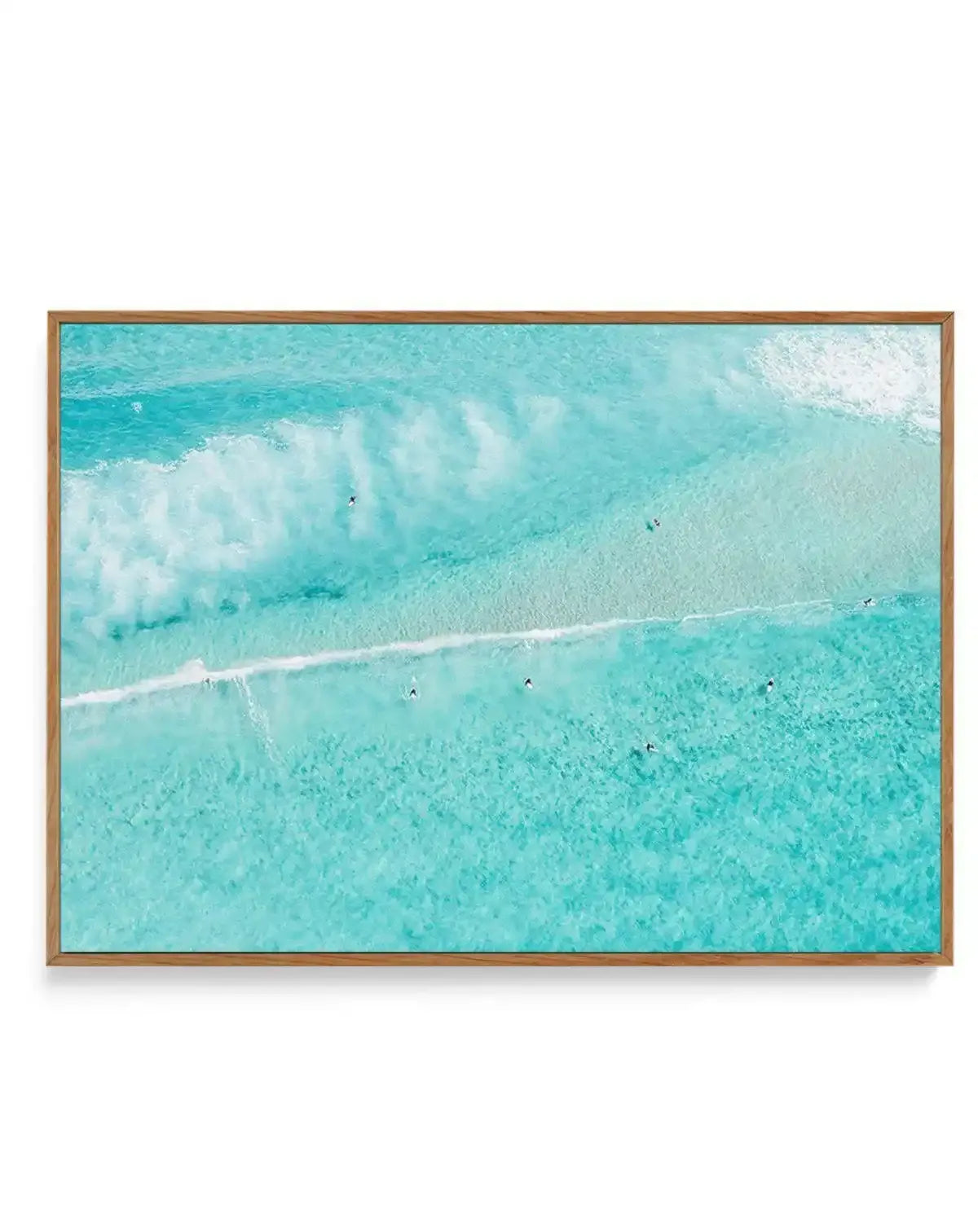 Aerial photograph of turquoise ocean water with white-capped waves rolling toward a sandy beach. Small surfers dot the crystalline shallow waters in various shades of aqua and teal. The image captures multiple wave breaks and a white sandy shoreline. Printed on canvas with a natural wood frame and white border.