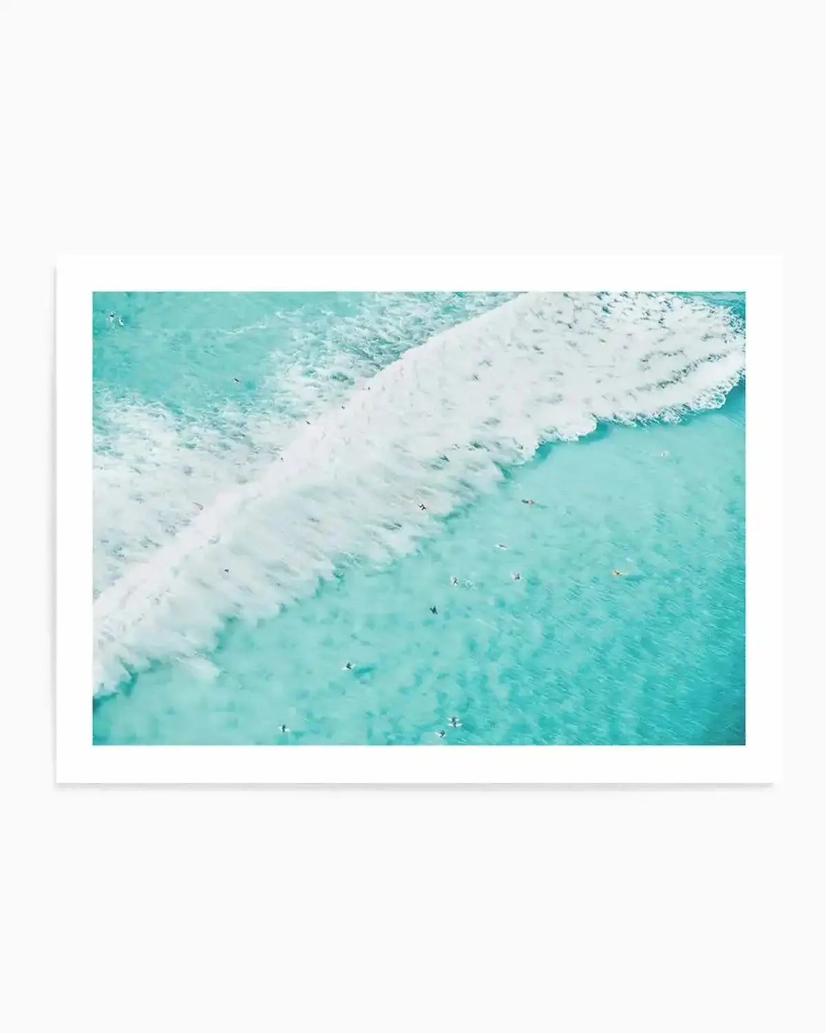 Aerial photography art print of a turquoise ocean with a large white wave breaking diagonally across the frame. Numerous small dark figures, likely surfers, are scattered in the water, some near the wave and others further out. The water transitions from a lighter, foamy turquoise near the wave to a deeper, clearer turquoise further from the shore.