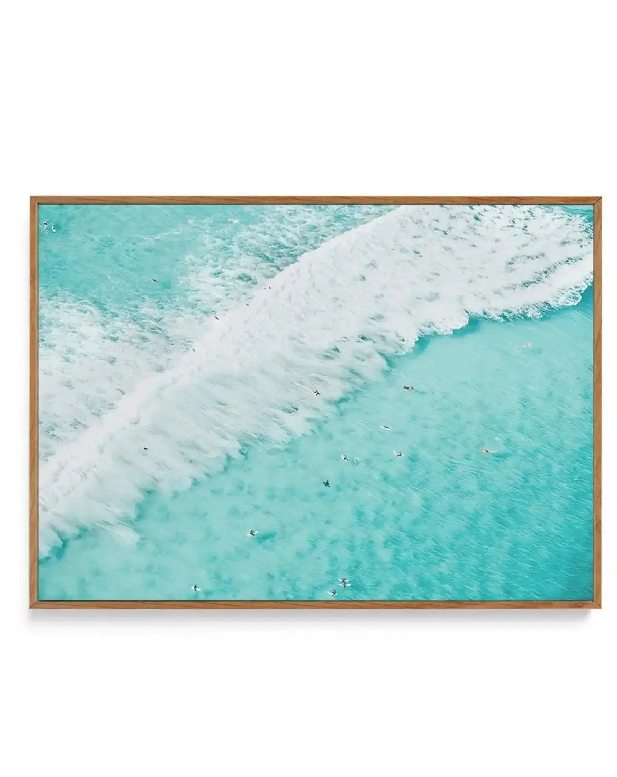 Aerial photograph of turquoise ocean water with white foamy waves rolling diagonally across the frame. Several small surfers in wetsuits are visible as dark specks in the water. The image is printed on canvas and framed in a warm natural wood frame with a subtle border.