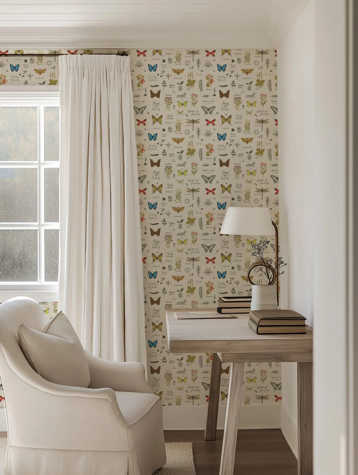 Lifestyle shot of a cozy reading nook featuring the Book of Butterflies wallpaper. The wallpaper has a repeating pattern of various butterflies, moths, dragonflies, and botanical illustrations in muted tones of blue, red, yellow, and green on a light cream background. The room includes a white armchair with a throw pillow, a wooden desk with books and a lamp, and a large window with white curtains.