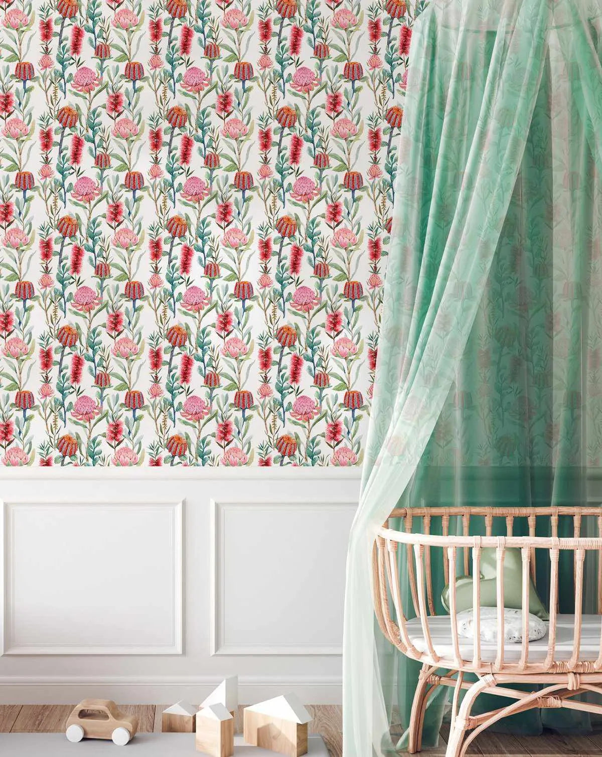 Lifestyle shot of a nursery featuring botanical garden wallpaper with a repeating pattern of various Australian native flowers in shades of pink, red, and orange with green foliage on a white background. The wallpaper is installed above white wainscoting. A light wood crib with a sheer mint green canopy is on the right, and wooden toys are on the floor in the foreground.