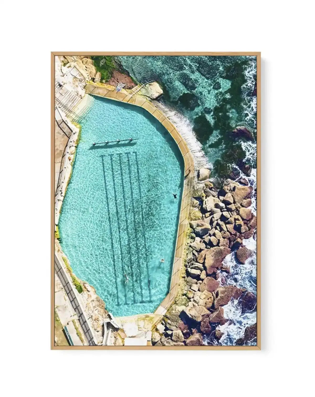 Aerial photography canvas print of the Bronte Ocean Pool in Sydney, Australia, displayed in a natural wood frame. The pool is a vibrant turquoise, with visible lane lines and a few swimmers, surrounded by a concrete edge. Beyond the pool, the ocean transitions from clear, shallow turquoise water over rocks to deeper, darker blue-green water with white foamy waves crashing against the rocky coastline. The composition captures the contrast between the man-made pool and the natural rugged coastline.