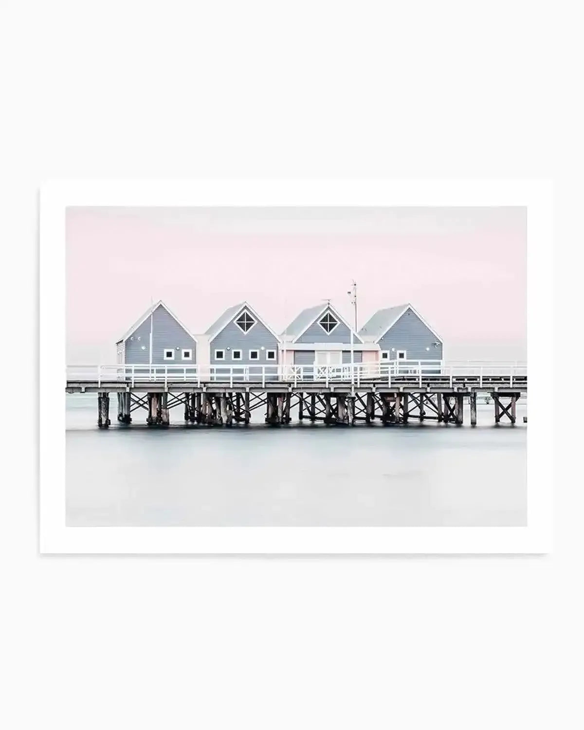 Framed photograph of the Busselton Jetty in Western Australia, featuring a row of four small, light grey wooden beach houses with white trim and white picket fences, standing on a long wooden pier over calm, pale blue water. The sky above is a soft, pastel pink, creating a serene and minimalist coastal scene.