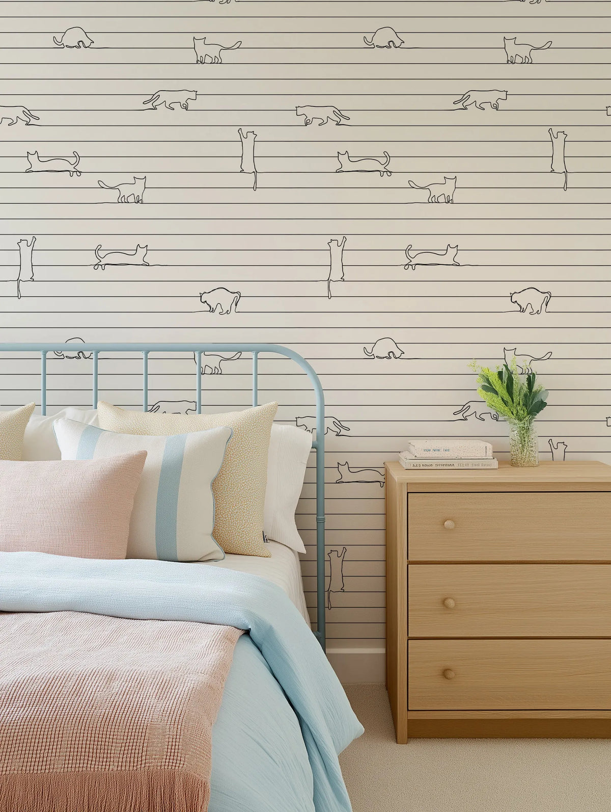 "Bedroom featuring wallpaper with a repeating pattern of continuous line art cats in various poses on a cream background with thin horizontal stripes. The wallpaper is displayed behind a light blue metal bed frame with a bed dressed in light blue and pink bedding, and a light wood bedside table with a vase of greenery and books."