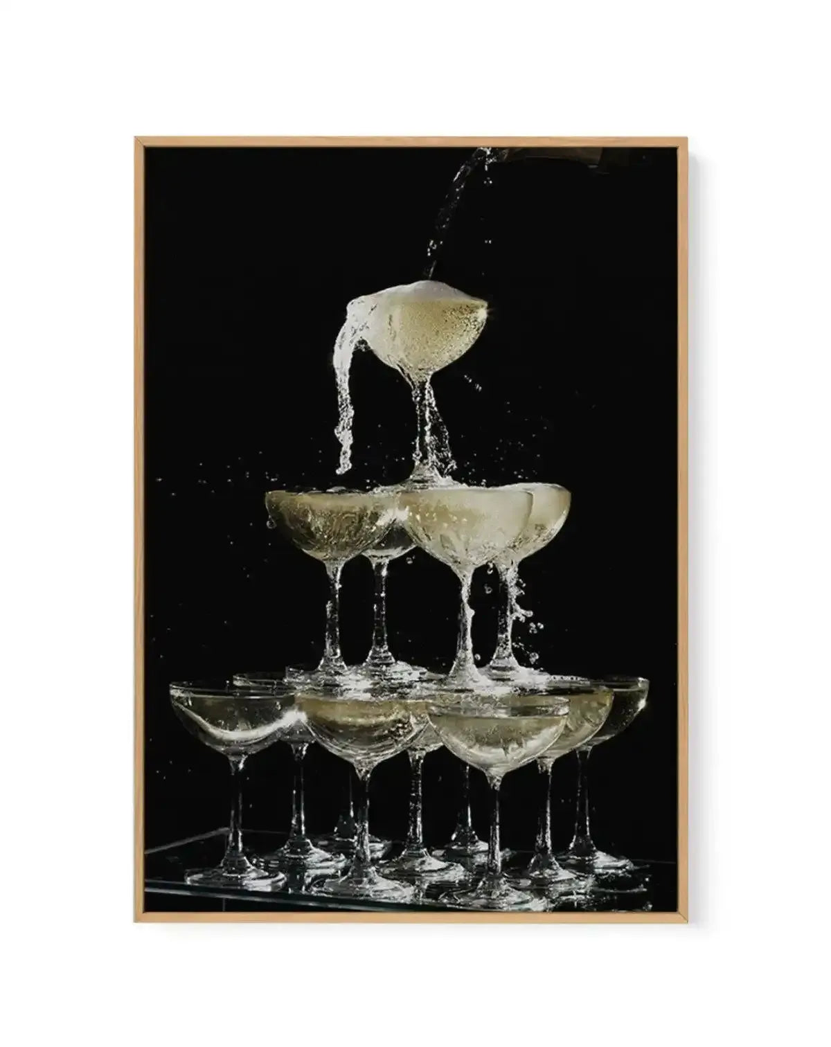 Framed canvas art print featuring a high-speed photograph of champagne glasses stacked in a pyramid formation against a black background. Water and champagne splash dramatically upward from the top glass, captured mid-motion with crystalline detail. The glasses contain pale golden liquid and are arranged in three tiers with approximately five glasses at the base. The image has a cream-colored wooden frame with a white border.
