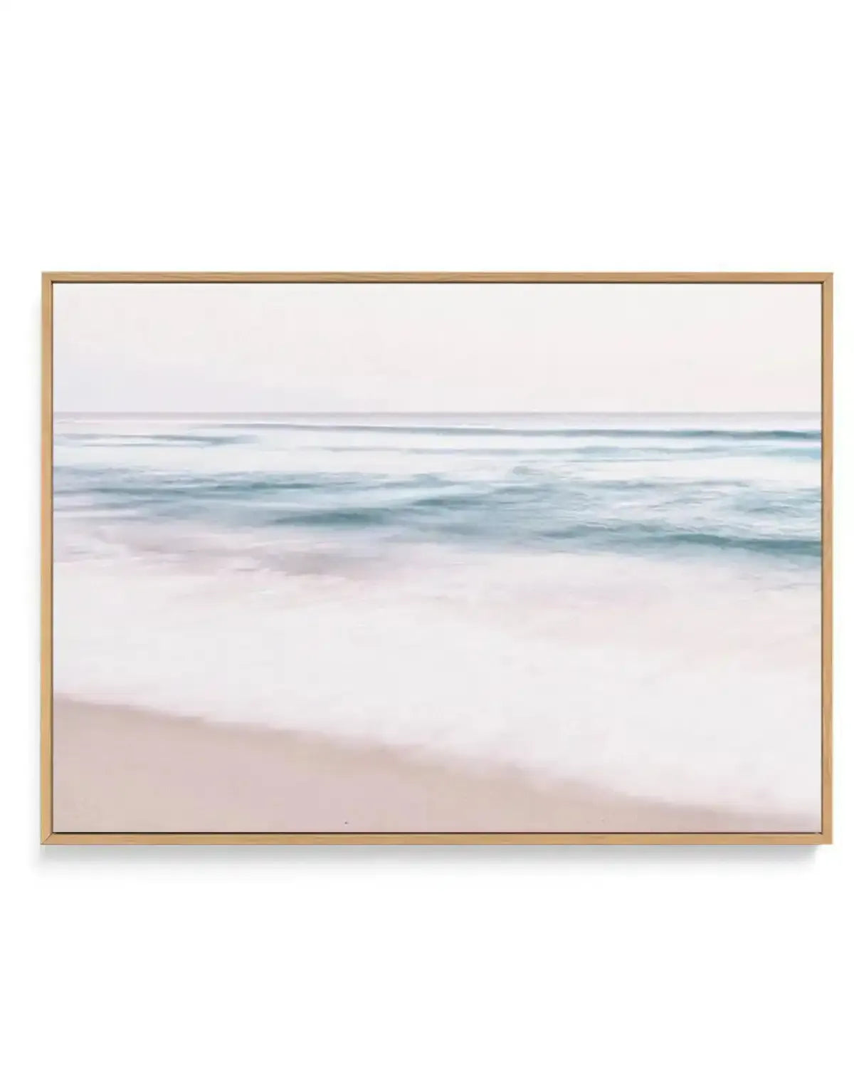 Framed canvas art print featuring a soft, blurred seascape with pale sandy beach in the foreground transitioning to gentle ocean waves in shades of seafoam green and slate blue, beneath a soft cream sky. The photograph has a dreamy, motion-blur quality. The canvas is mounted in a natural wood frame with a white border.