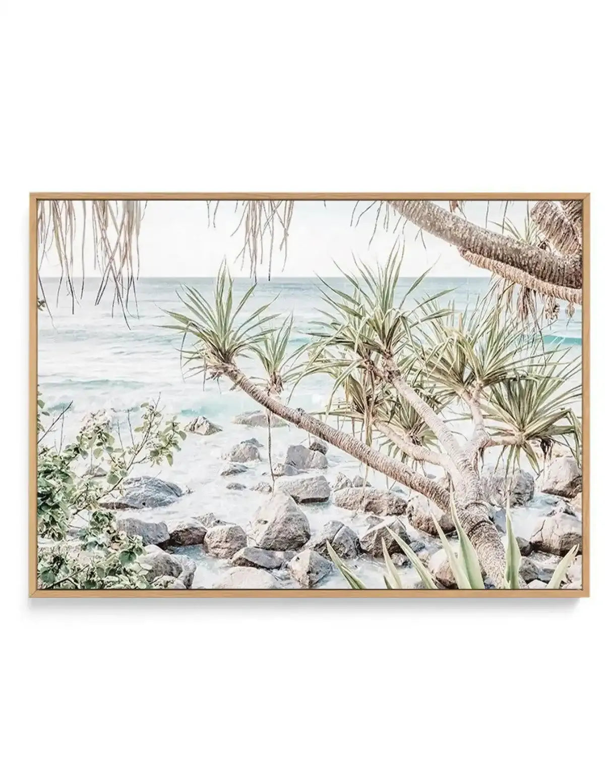 Framed canvas print of a serene coastal beach scene with a natural wood frame. The photograph captures light blue ocean water gently lapping against a rocky shore with white sand. Lush green tropical plants with long, spiky leaves and gnarled branches frame the foreground, creating a tranquil, natural composition.