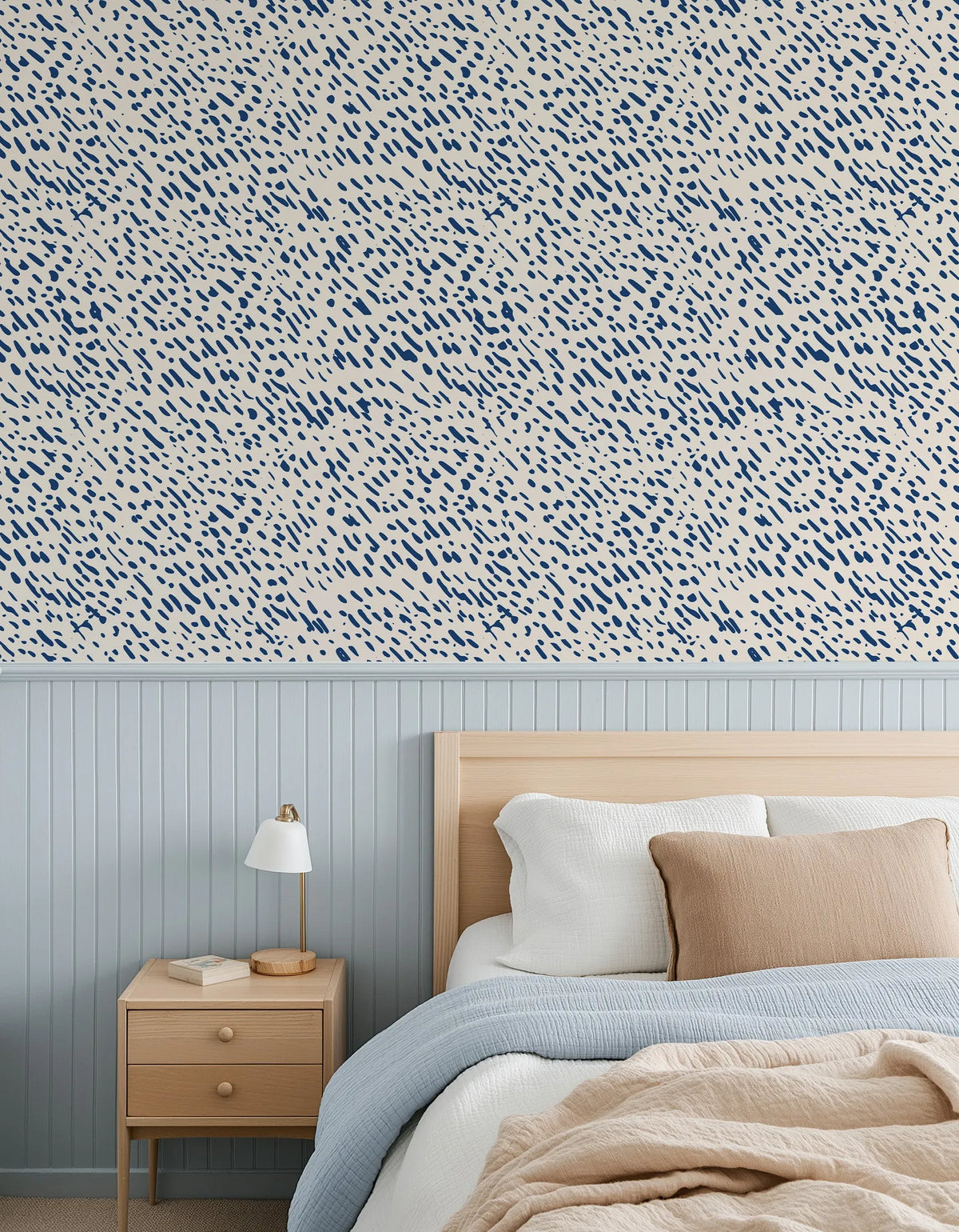"Bedroom featuring Dapple Wallpaper with a repeating pattern of small, hand-painted dash marks in deep navy blue on a warm cream background, creating a textured, organic look. The wallpaper is displayed above light blue wainscoting and a natural wood bed frame with white, light blue, and beige bedding, and a wooden bedside table with a white lamp."