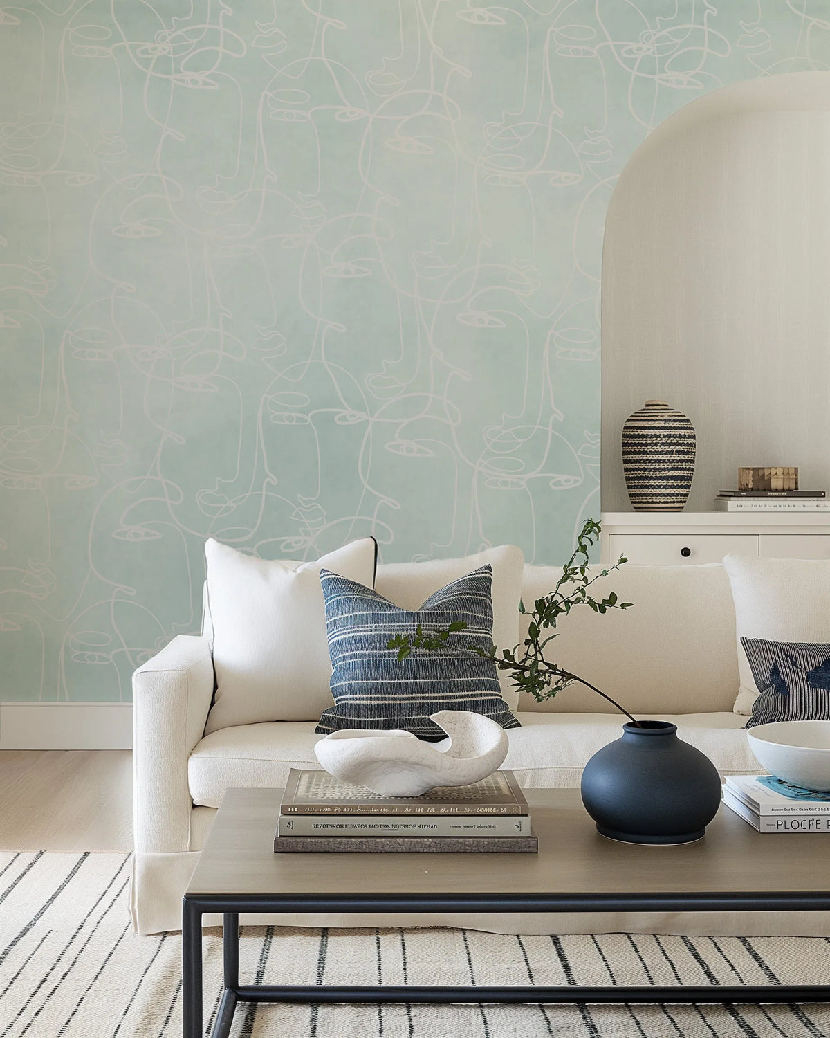 Lifestyle shot of a minimalist living room featuring peel and stick wallpaper with a repeating pattern of abstract line art faces in white on a seafoam green background. The wallpaper covers a large wall behind a white sofa with blue and white striped cushions. A light wood coffee table with books and decorative objects sits in front of the sofa.
