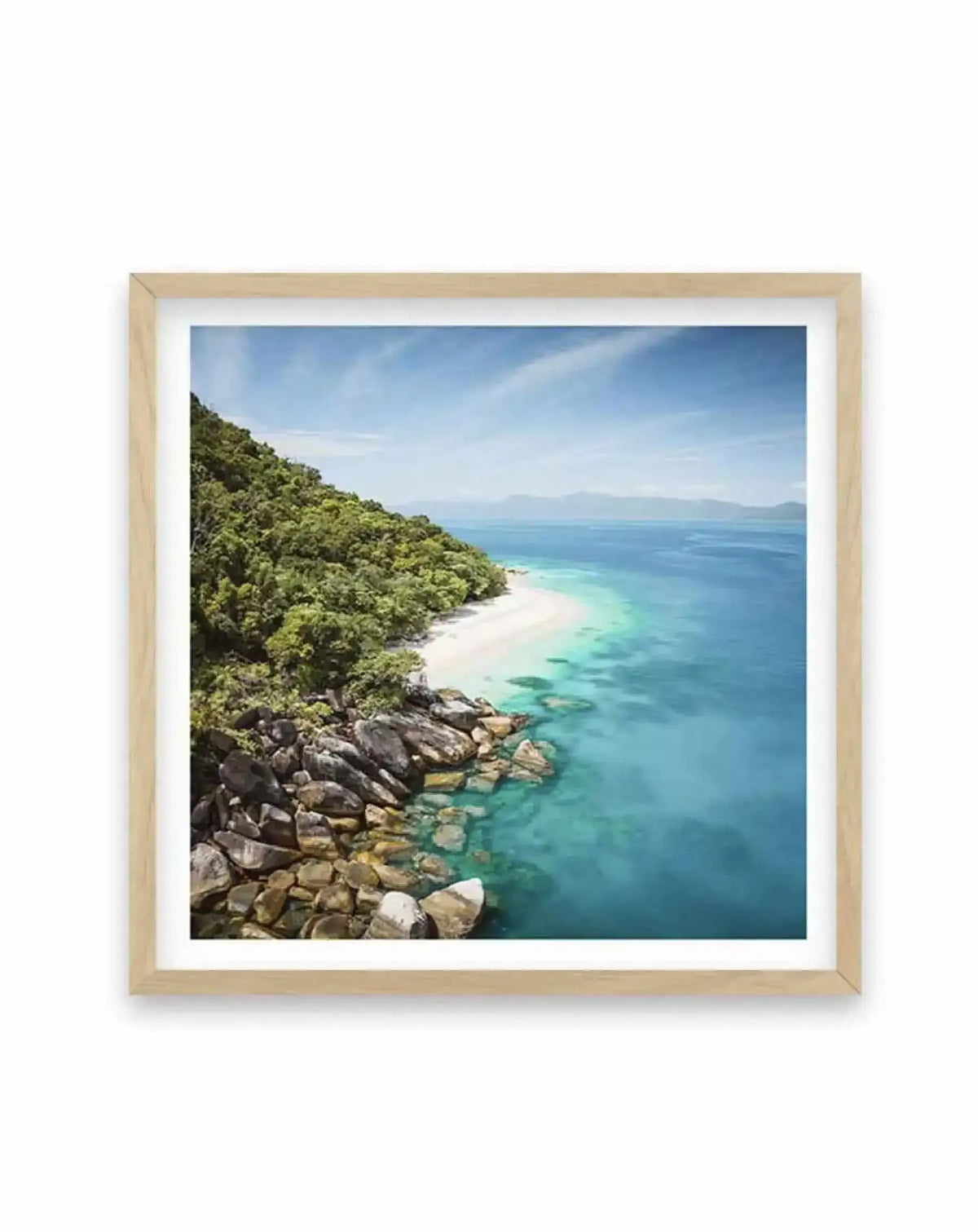 Framed photograph of a tropical beach scene with a natural wood frame and white border. The composition features a lush green island hillside sloping down to a crescent of white sand beach, bordered by clear turquoise and deep blue ocean under a bright blue sky with wispy clouds. Large grey rocks are visible along the shoreline where the water meets the land.