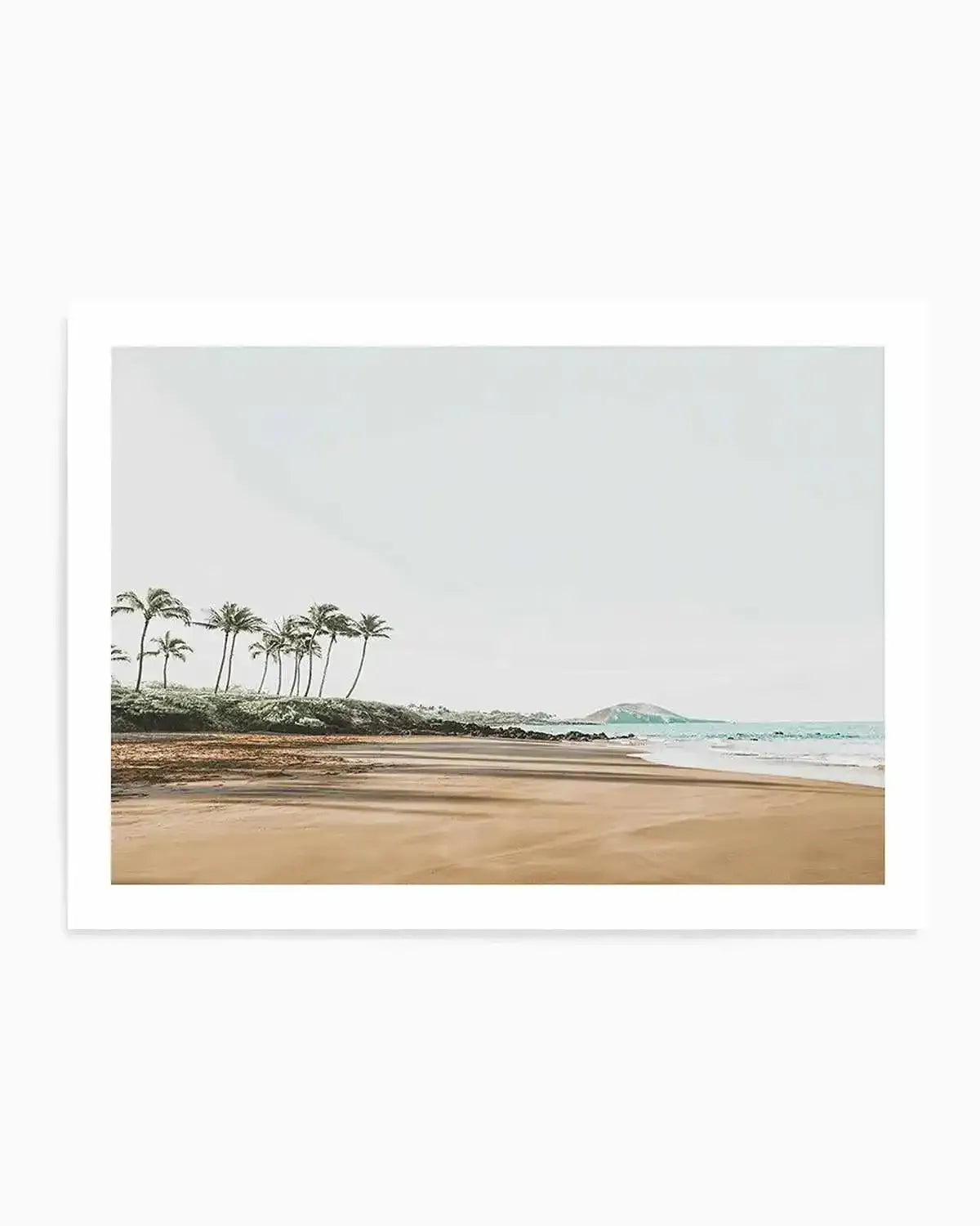 Framed photograph of a serene coastal beach scene with a light, airy aesthetic. The composition features a line of palm trees on the left, casting long shadows across the light brown sand. Gentle waves with a light blue hue meet the shore on the right, and a small island or hill is visible in the distance under a pale, overcast sky. The print is displayed in a white frame with a white border.