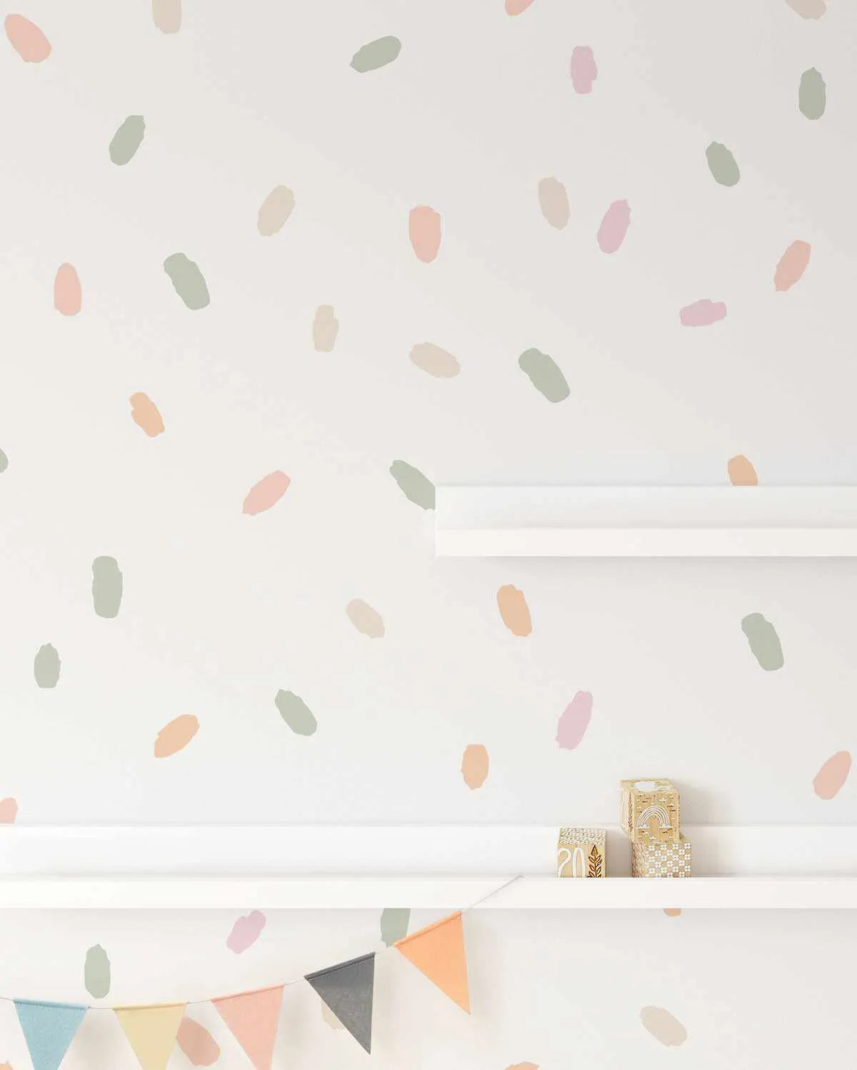 Wall Decal:
"Peel and stick wall decals featuring 174 irregular, hand-painted dots in a mix of pastel shades including dusty pink, sage green, peach, and beige, scattered across a white wall. The dots vary slightly in size and shape, resembling brushstrokes or confetti, creating a playful, whimsical pattern. Two white shelves are visible, with a small decorative block and a gold box on the lower shelf, and a string of pastel-colored bunting hanging below."