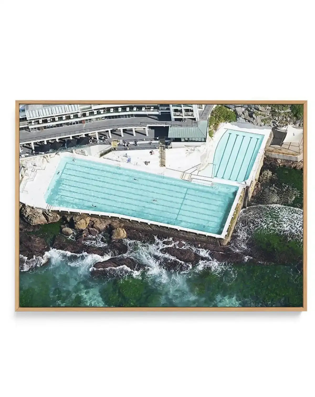 "Gallery-wrapped canvas print of an aerial photograph of the Bondi Icebergs pool in Sydney, Australia, with a natural wood frame. The image captures the iconic ocean pool with its bright turquoise water and lane lines, nestled against the rocky coastline with crashing waves and deep green ocean water. In the background, the Bondi Icebergs Club building with its various levels and white structures is visible."