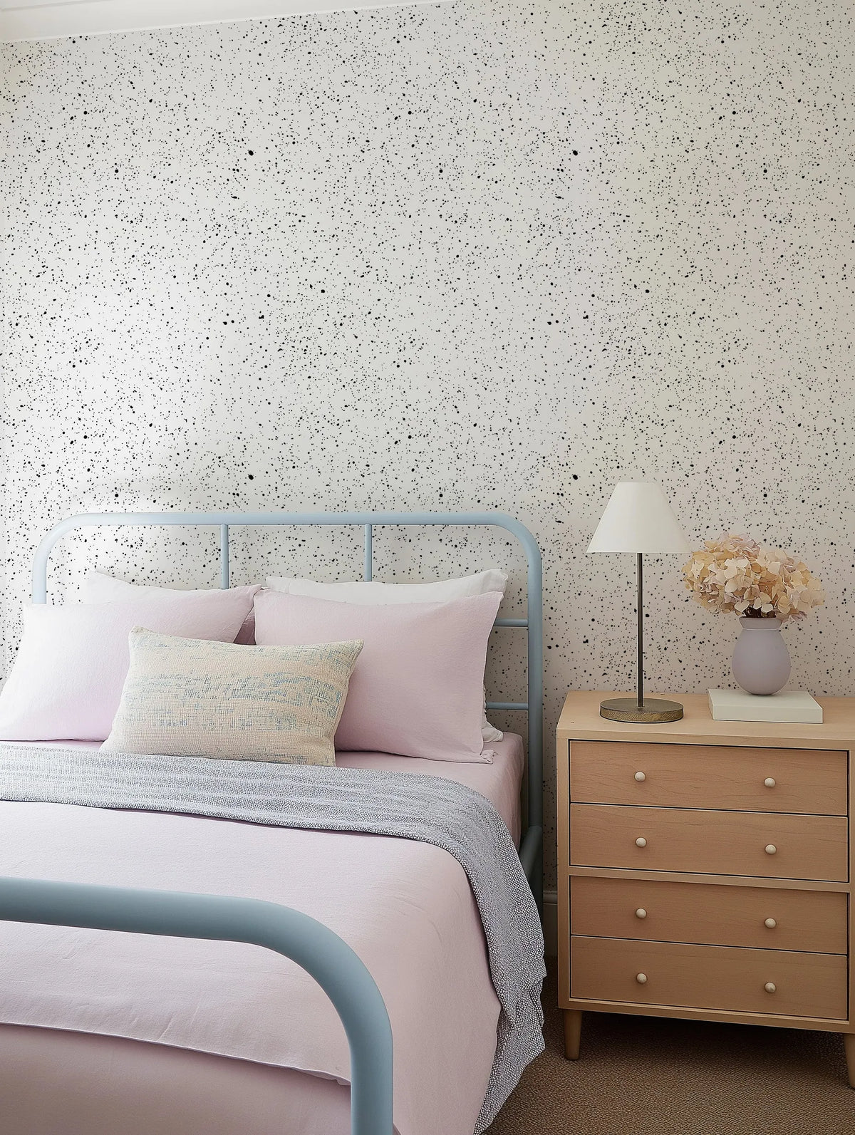 A bedroom featuring a wall covered in "Ink Spots Wallpaper" with a light background and scattered black speckles of varying sizes, creating a playful, abstract pattern. A light blue metal bed frame with pink bedding and pillows is positioned against the wallpapered wall. A textured cream and light blue throw pillow rests on the bed. To the right, a light wood nightstand holds a brass-based lamp with a cream shade and a light grey vase filled with dried, light pink hydrangeas.