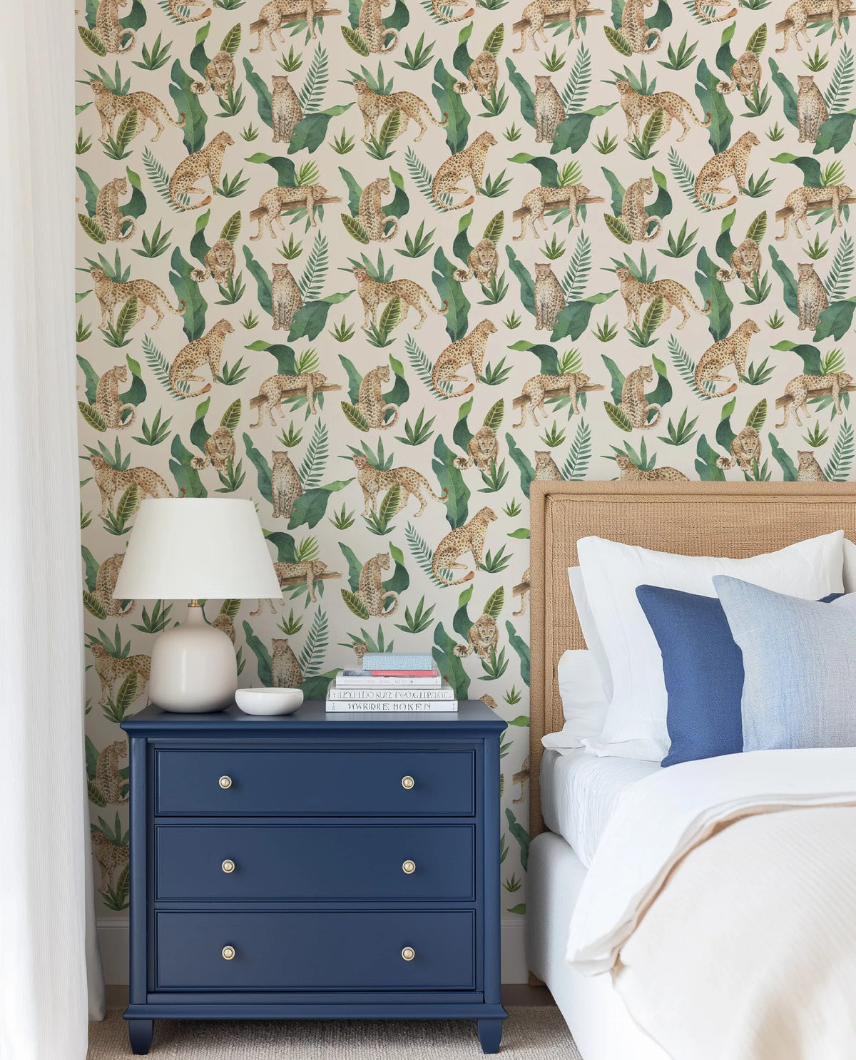 Lifestyle shot of a bedroom featuring Jungle Leopards wallpaper behind a bed with a woven rattan headboard. The wallpaper has a repeating pattern of hand-painted leopards in various poses, surrounded by lush green tropical leaves on a light cream background. The room includes a navy blue bedside table with a white lamp, and the bed is dressed with white and blue pillows and white bedding.