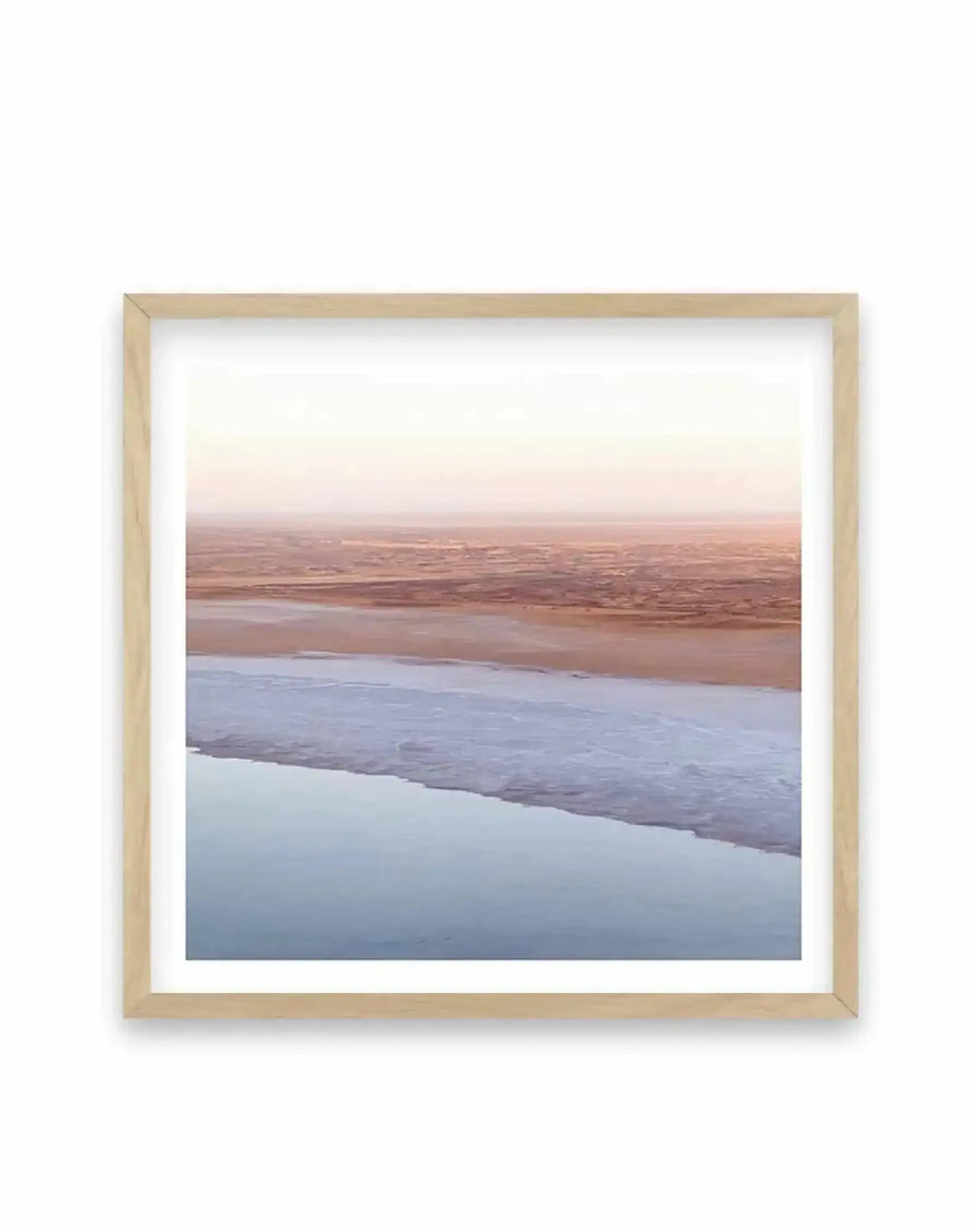 Framed photography art print of a vast, flat landscape with a body of water in the foreground, displayed in a natural oak frame with white border. The scene depicts a pale blue body of water meeting a light brown, sandy shore, which stretches into the distance under a soft, hazy sky in shades of pale pink and cream.