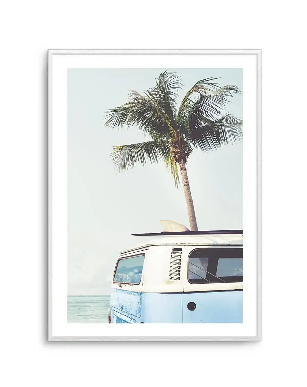 Framed photography art print of a light blue and white vintage Kombi van with a surfboard on its roof, parked on a beach next to the ocean, with a tall palm tree rising behind it against a pale blue sky, displayed in a white frame with a white border.