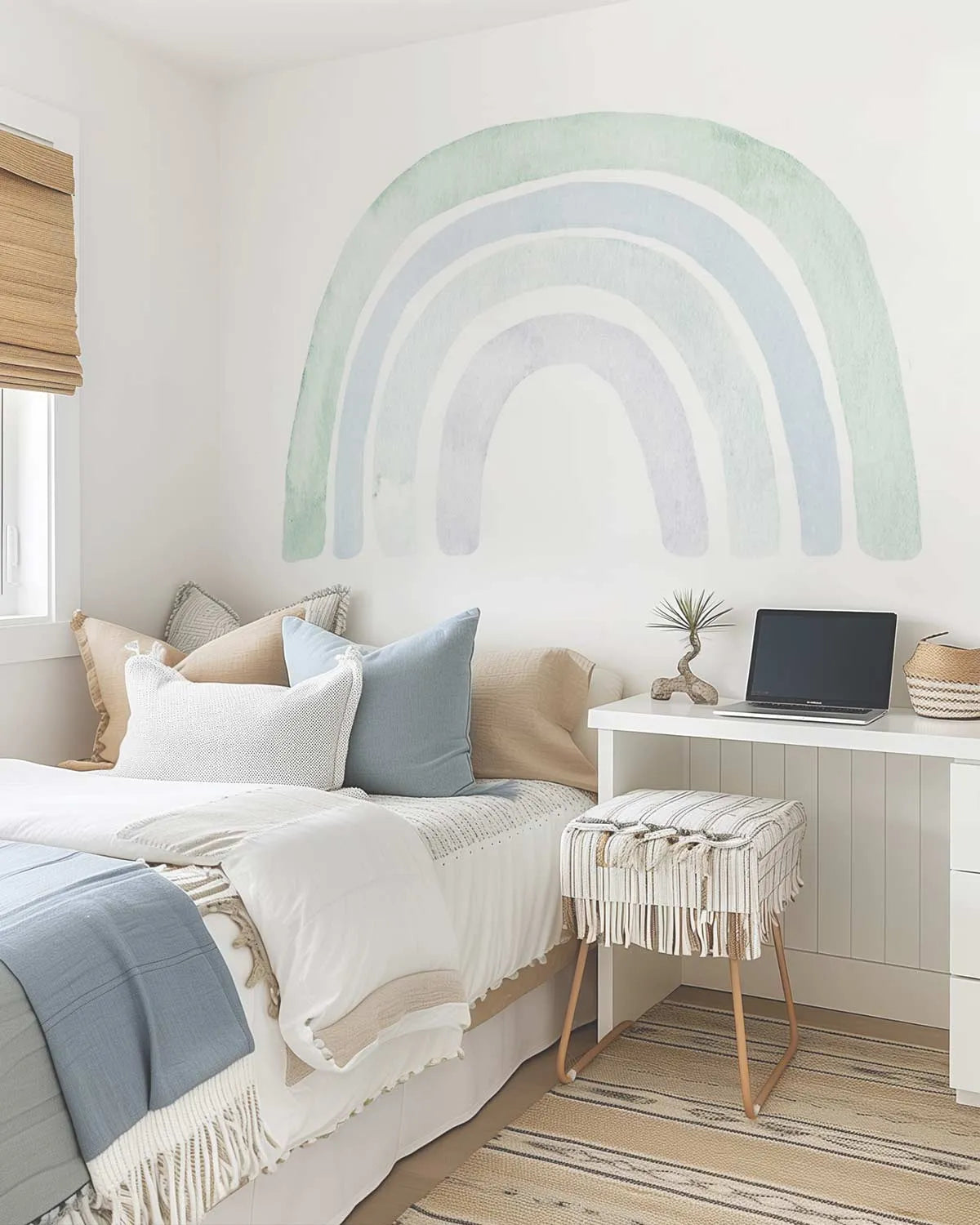 Lifestyle shot of a bedroom featuring a large peel and stick wall decal of a rainbow in muted pastel shades of sage green, light blue, and pale grey, painted with a soft watercolor effect. The decal is applied to a white wall above a bed dressed with white, cream, and light blue bedding, and a small white desk with a laptop and a striped stool.