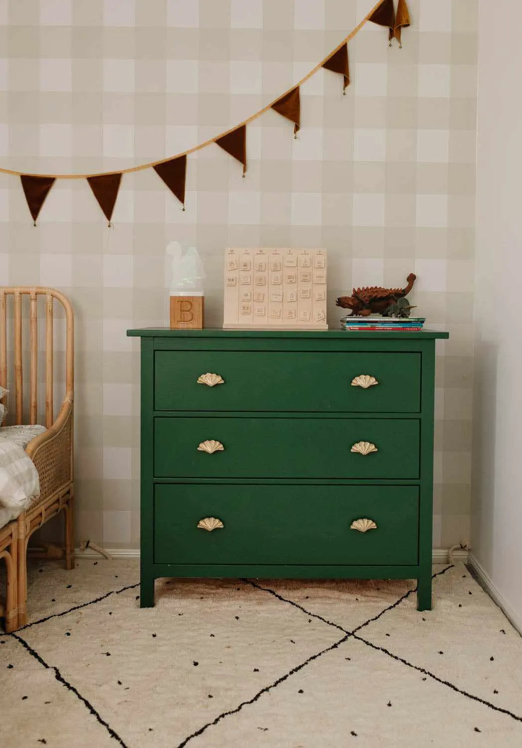 Lifestyle shot of a child's bedroom featuring a large gingham check wallpaper in a new neutral green and off-white pattern. The wallpaper serves as a backdrop for a dark green dresser with gold shell-shaped pulls, a rattan crib, and a cream rug with black geometric lines and speckles. The dresser is adorned with a wooden block calendar, a light-up "B" initial, and a dinosaur toy on a stack of books.