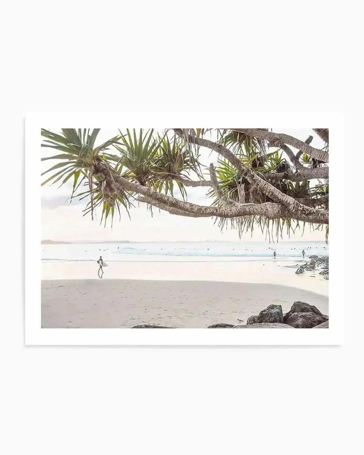 Framed photograph of a coastal beach scene with a natural wood frame and white border. The composition features a surfer walking along wet sand at low tide with gentle waves and an overcast sky stretching to the horizon, framed by the branches of a pandanus tree in the foreground.