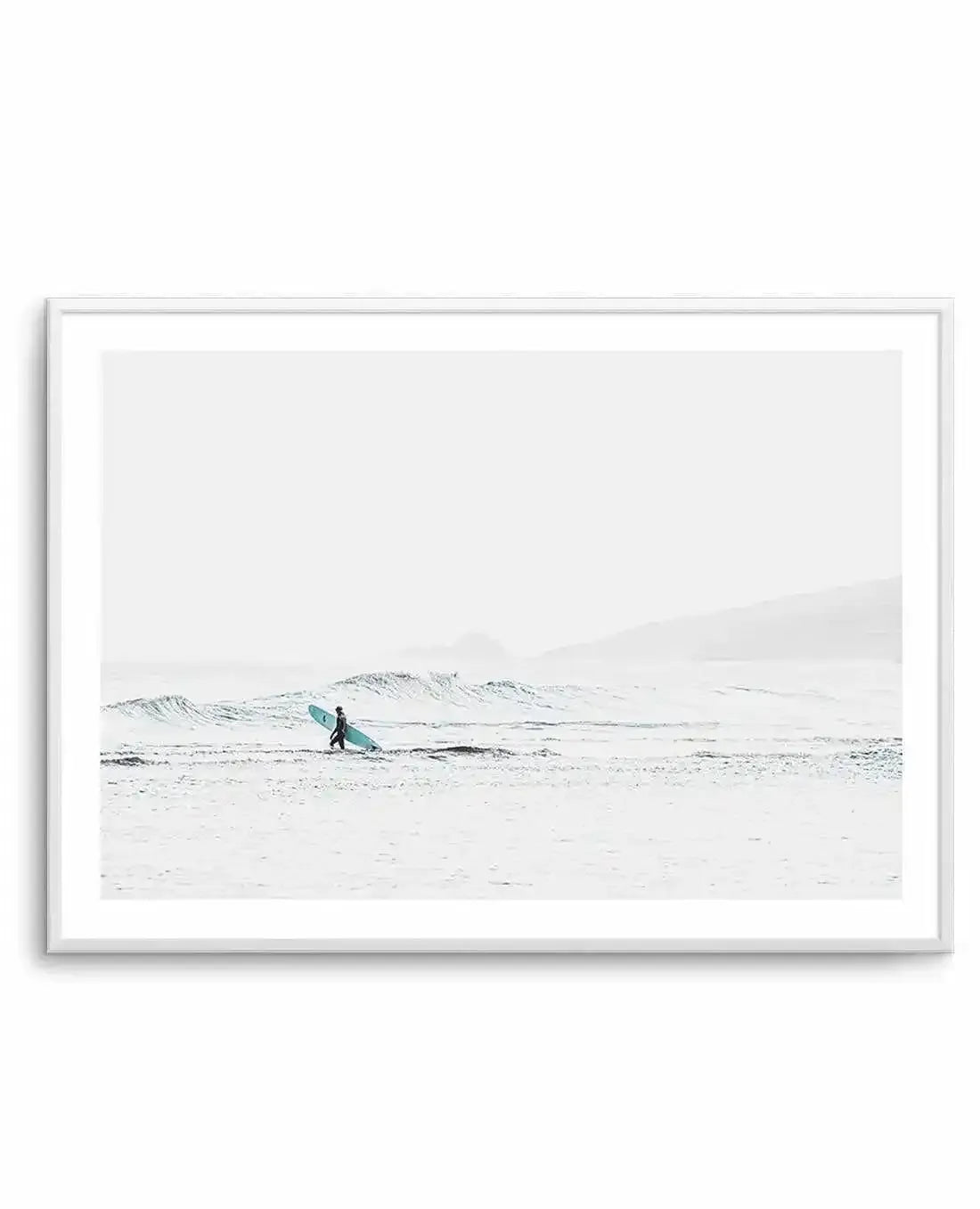 Framed black and white photograph of a coastal beach scene with a white frame and white border. The composition features a lone surfer walking along wet sand at low tide with gentle waves and an overcast sky stretching to the horizon.