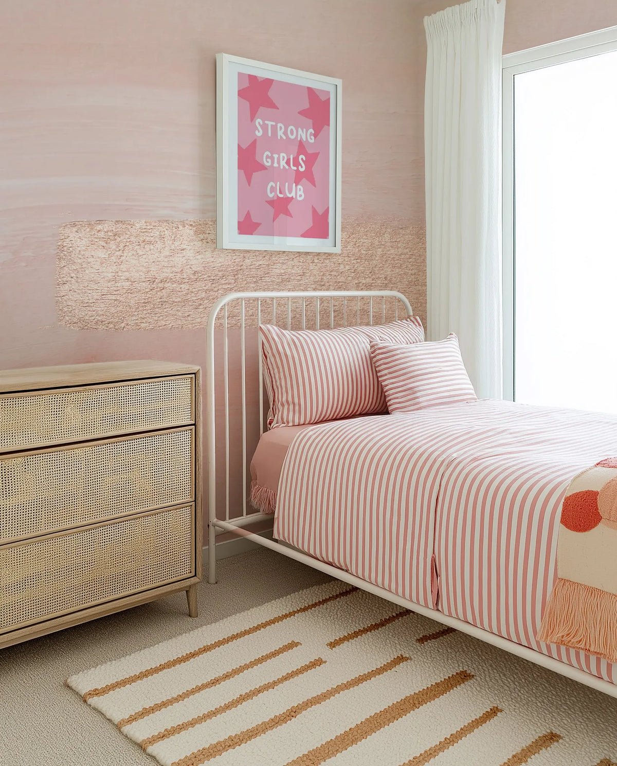 "Peel and stick wallpaper featuring a horizontal ombre design with a soft blush pink at the top fading into a textured, lighter pink with horizontal brushstrokes at the bottom. The wallpaper is displayed in a child's bedroom with a white metal bed frame, pink and white striped bedding, a rattan dresser, and a white rug with orange stripes."