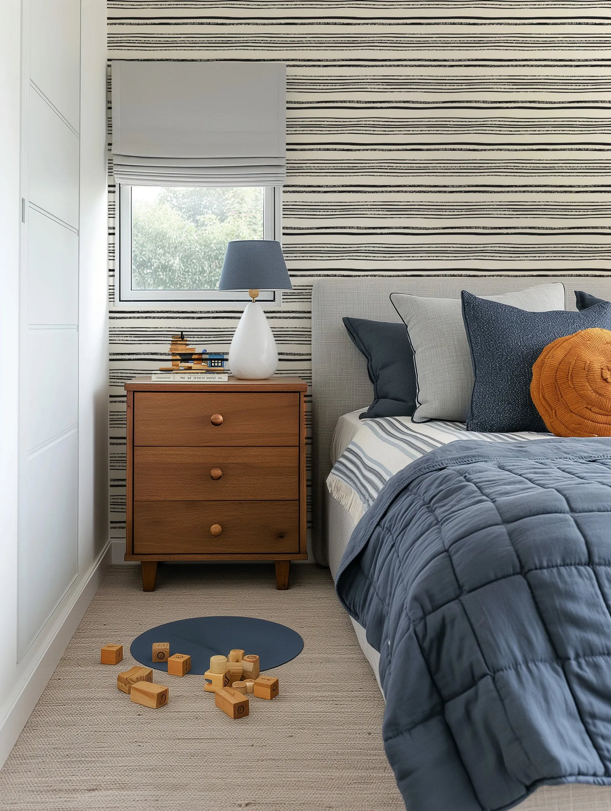 Lifestyle shot of a child's bedroom featuring a wallpaper with a repeating pattern of hand-drawn horizontal stripes in black on a cream background. The stripes vary in thickness and texture, some appearing solid and others with a distressed, brushstroke quality. The wallpaper covers the wall behind a bed with a light grey upholstered headboard and a wooden nightstand. The room includes a window with a light grey roman blind, a blue quilted duvet, and wooden toys on a light-coloured rug.
