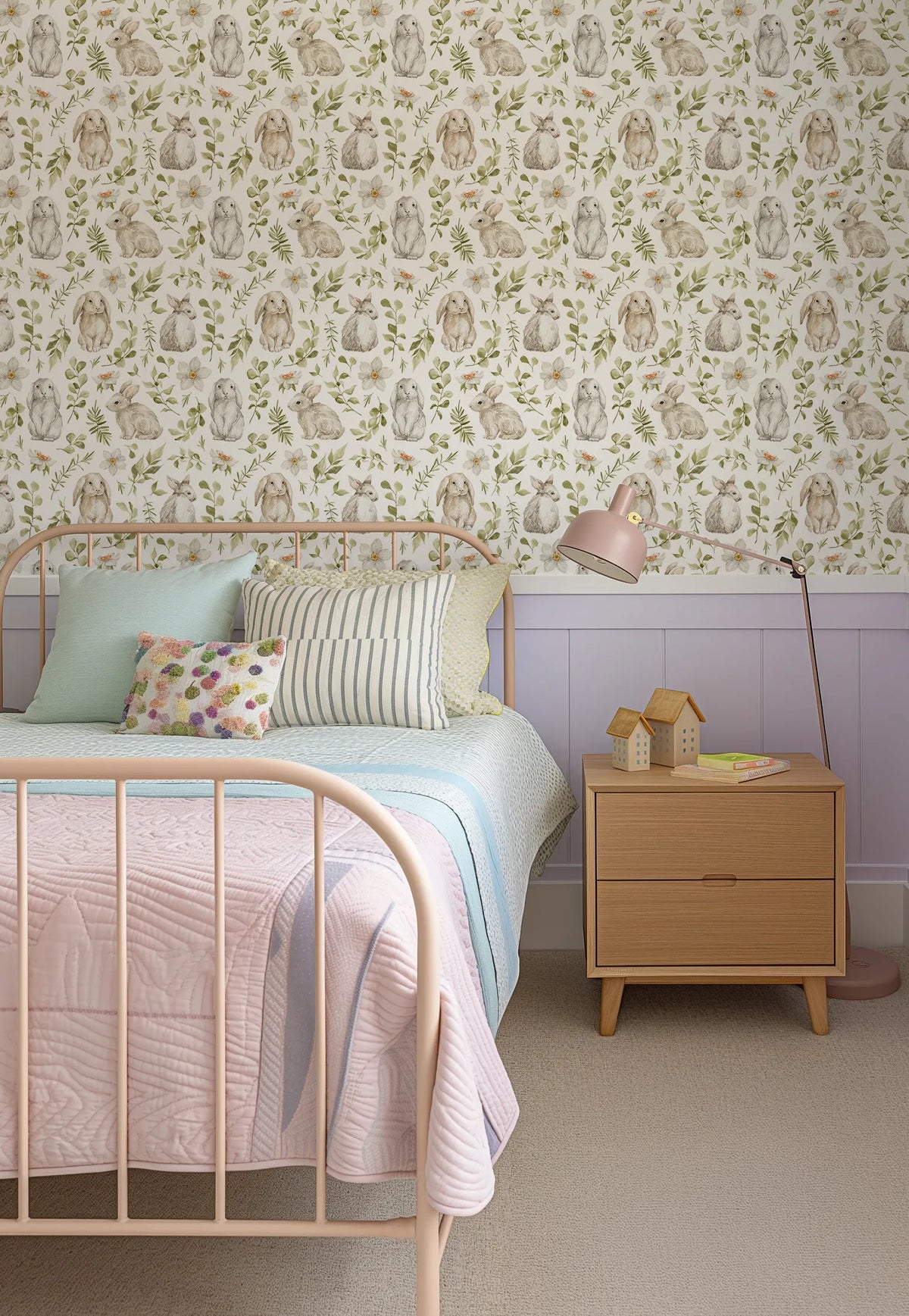 "Little Bunny wallpaper featuring a repeating pattern of hand-painted bunnies in various shades of brown and grey, interspersed with delicate green foliage and white flowers, on a cream background. The wallpaper is displayed in a child's bedroom above a light purple wainscoting, with a pink metal bed frame, light blue and pink bedding, and a wooden bedside table with a pink lamp and decorative house figurines."