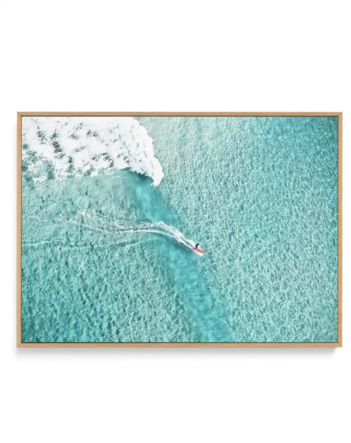 Framed aerial photograph of a surfer riding a wave in clear turquoise water, with a natural wood frame and white border. The composition captures the surfer on a red board, leaving a white foamy trail, with a large breaking wave to the left and clear, shallow water showing the sandy bottom.