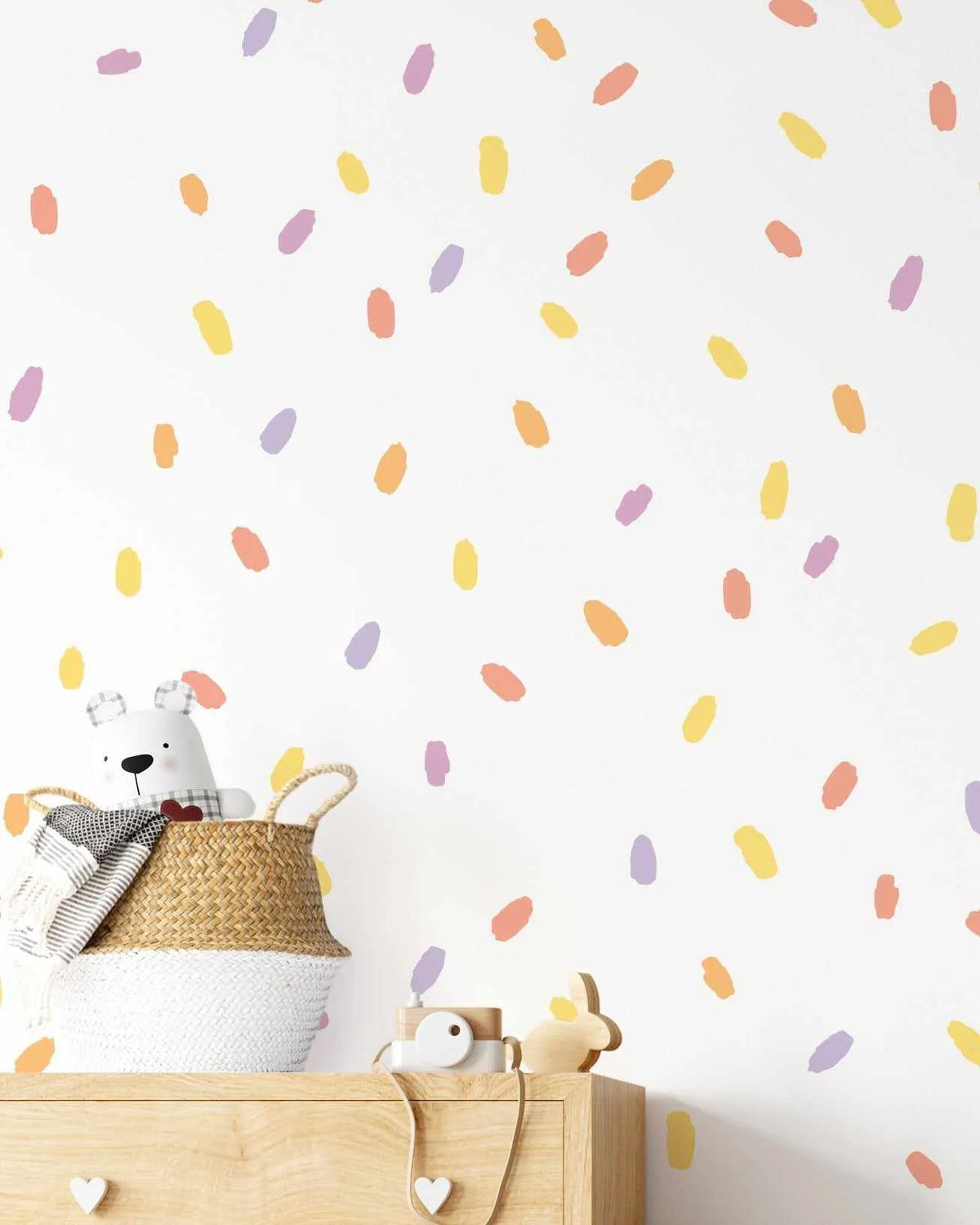 Nursery:
"White wall adorned with a random pattern of oval-shaped wall decals in pastel shades of yellow, orange, pink, and purple, creating a playful, confetti-like effect. Below, a woven basket holds a white teddy bear and a striped blanket, resting on a light wood dresser with heart-shaped handles and two wooden toys."