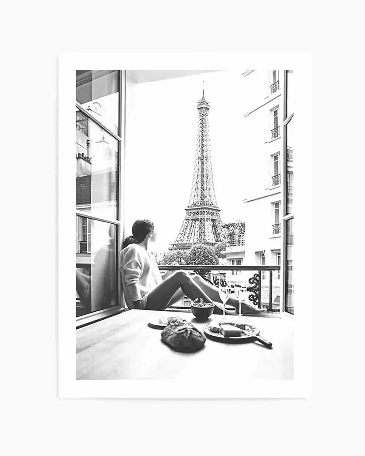 Framed black and white photograph of a woman sitting on a balcony overlooking the Eiffel Tower in Paris. The woman, seen from behind, wears a light-coloured top and looks out towards the iconic landmark. On the table in front of her are a loaf of bread, a bowl, and a plate with cheese and two wine glasses, suggesting a meal or picnic. The composition captures a serene moment with a classic Parisian view.