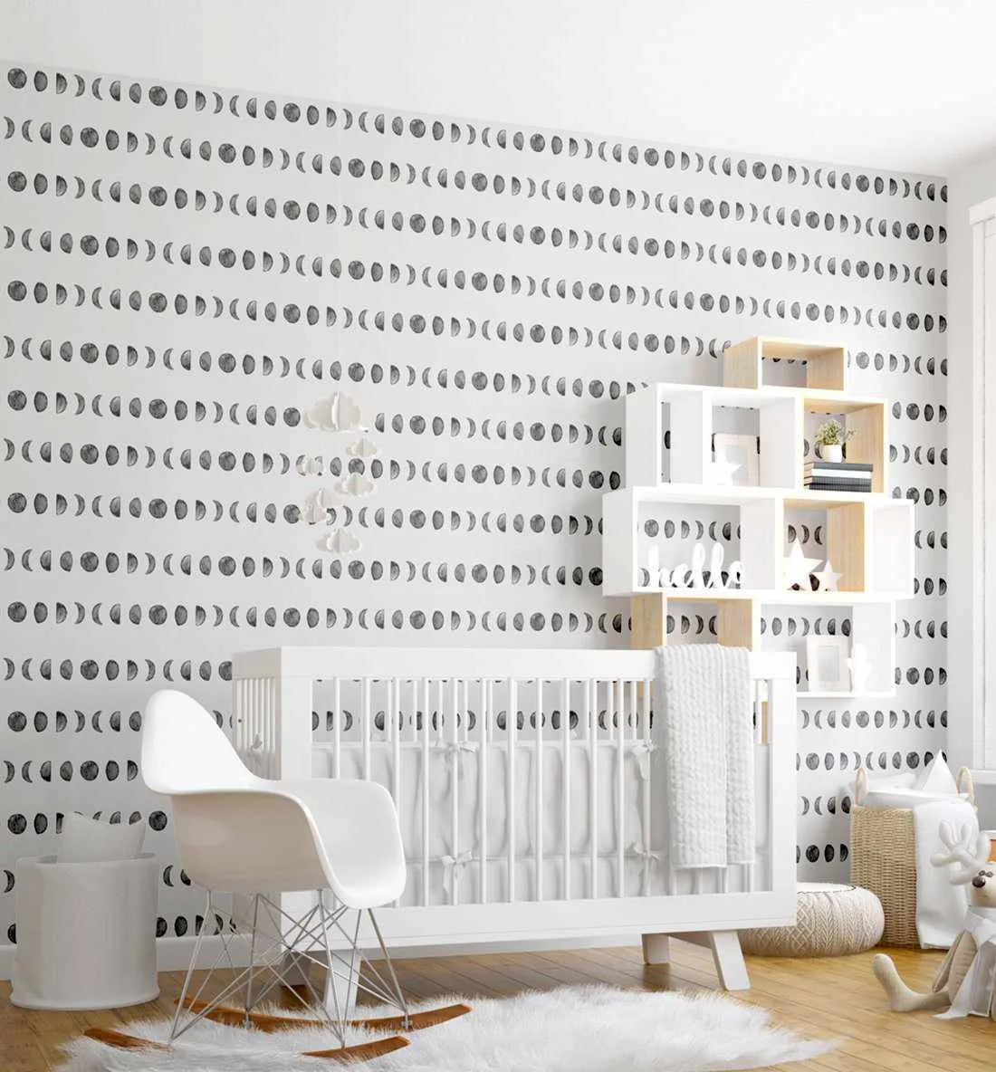Nursery:
"Moon Phases wallpaper displayed on the back wall of a modern nursery. The wallpaper features a repeating pattern of hand-drawn moon phases in charcoal grey on a crisp white background, arranged in horizontal rows. The room includes a white crib with white bedding, a white rocking chair, a white mobile with cloud shapes, a white and natural wood shelving unit with books and plants, a white fluffy rug, and a woven basket with a stuffed reindeer toy."