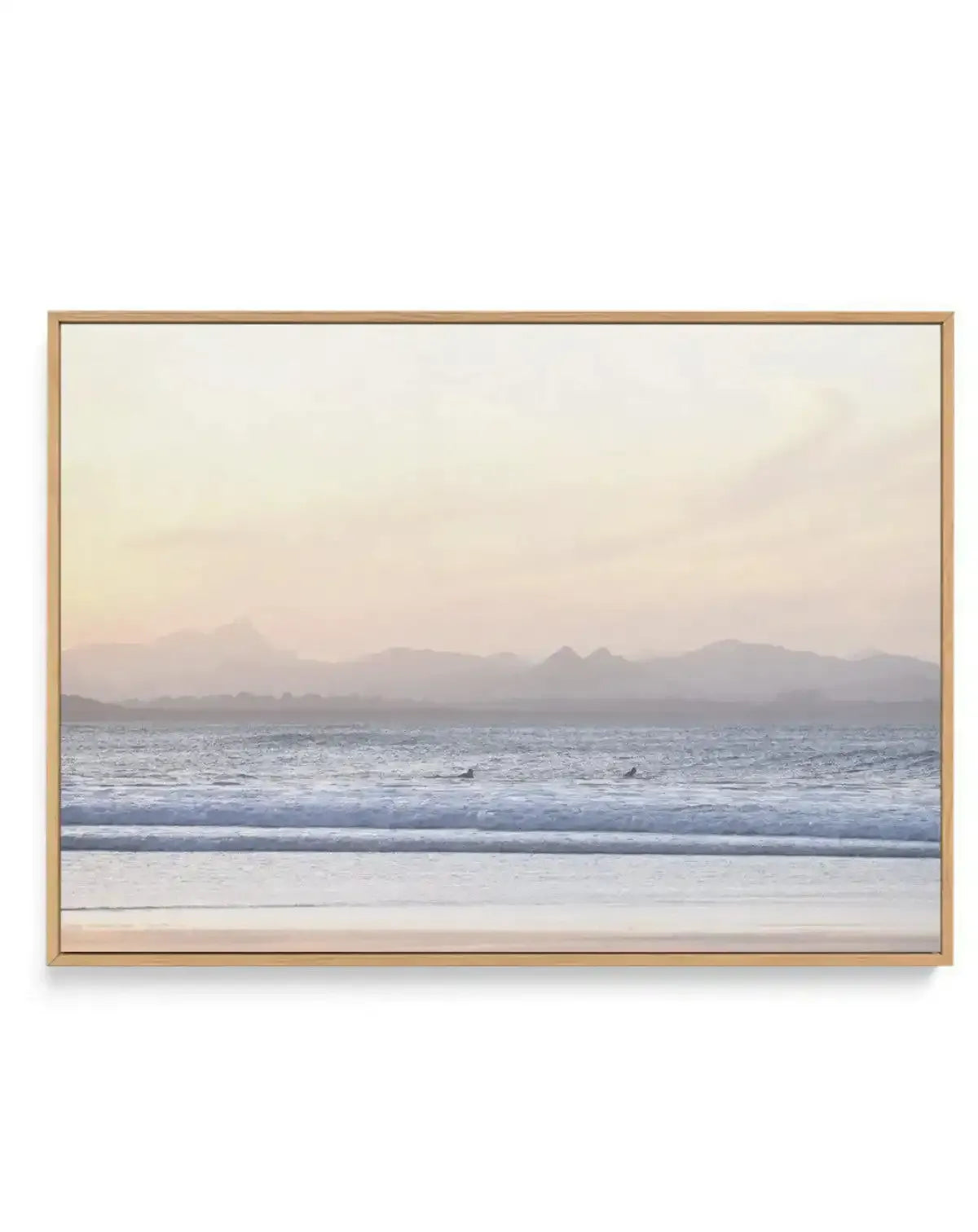 Framed canvas print of a serene coastal landscape at sunset, featuring a wide expanse of ocean with gentle waves breaking on the shore, and two surfers visible in the water. The sky is a soft gradient of pale yellow, peach, and light pink, transitioning to a hazy blue-grey over distant mountains. The foreground shows wet sand reflecting the sky, with the artwork displayed in a natural oak frame.