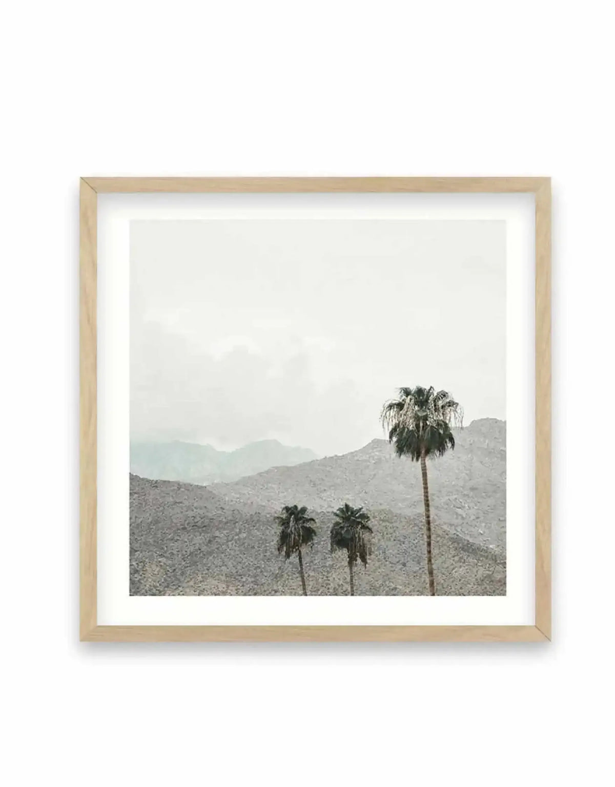 Framed square art print featuring a serene desert landscape photograph. Three palm trees with dark green fronds stand against rolling, rocky mountains rendered in muted grays and soft greens. Hazy mountains fade into a pale, overcast sky. The photograph has a soft, minimalist aesthetic with a dreamlike quality. Natural wood frame with white border surrounding the image.