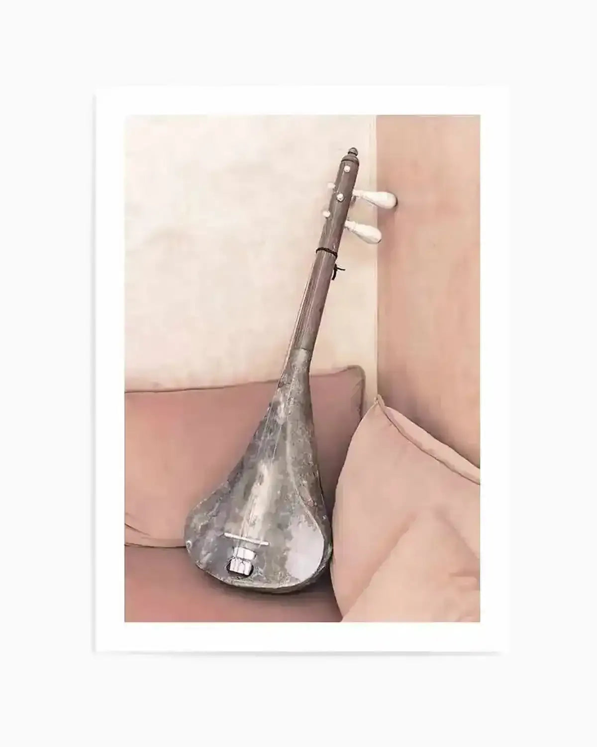 Framed art print featuring a photograph of a grey, pear-shaped stringed instrument with a long neck and white tuning pegs, leaning against a corner with light pink walls and cushions. The instrument has a metallic sheen and a small rectangular bridge near its base.