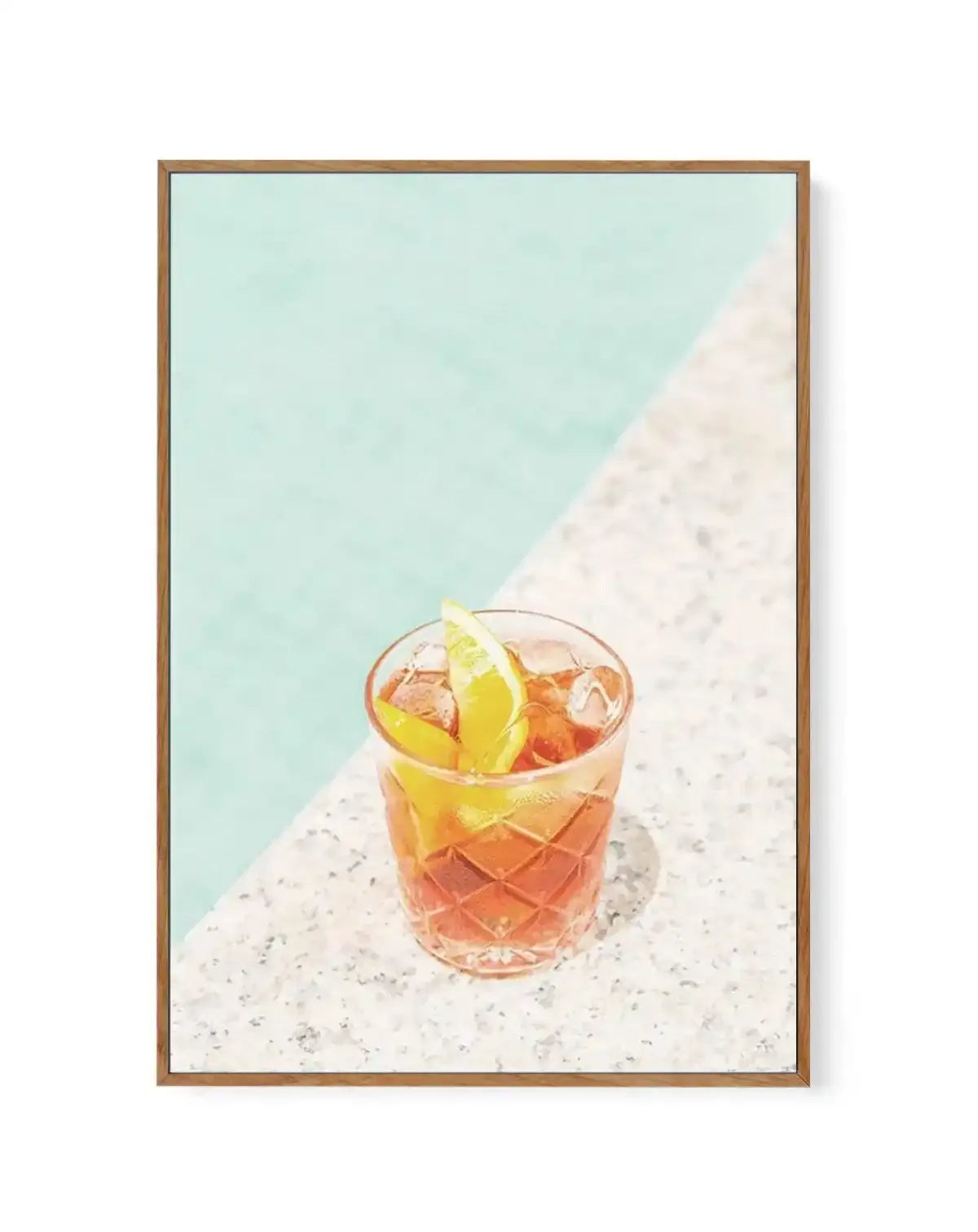 Framed canvas art print of a Negroni cocktail in a textured glass with ice and a yellow lime wedge, positioned on a speckled white surface against a turquoise water background and white sandy beach. The painting features soft watercolor tones in amber, coral, and citrine yellow. Natural wood-toned frame with white border surrounding the artwork.