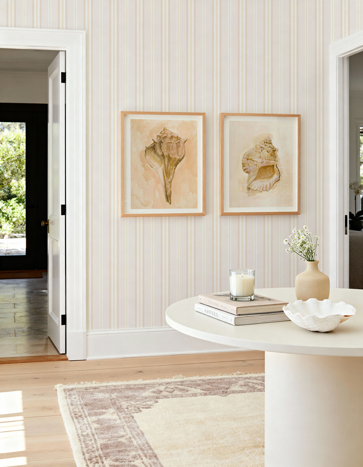 Lifestyle shot featuring Noosa Stripe Wallpaper in a bright entryway with two shell art prints and a round white table. The wallpaper has vertical stripes in soft cream, pale yellow, and light peach, creating a subtle, warm pattern. The room includes a light wood floor, a cream and brown patterned rug, and a white door leading to a sunlit outdoor area with lush greenery. On the table, there's a lit candle, a stack of books, a small vase with white flowers, and a large white decorative shell bowl.