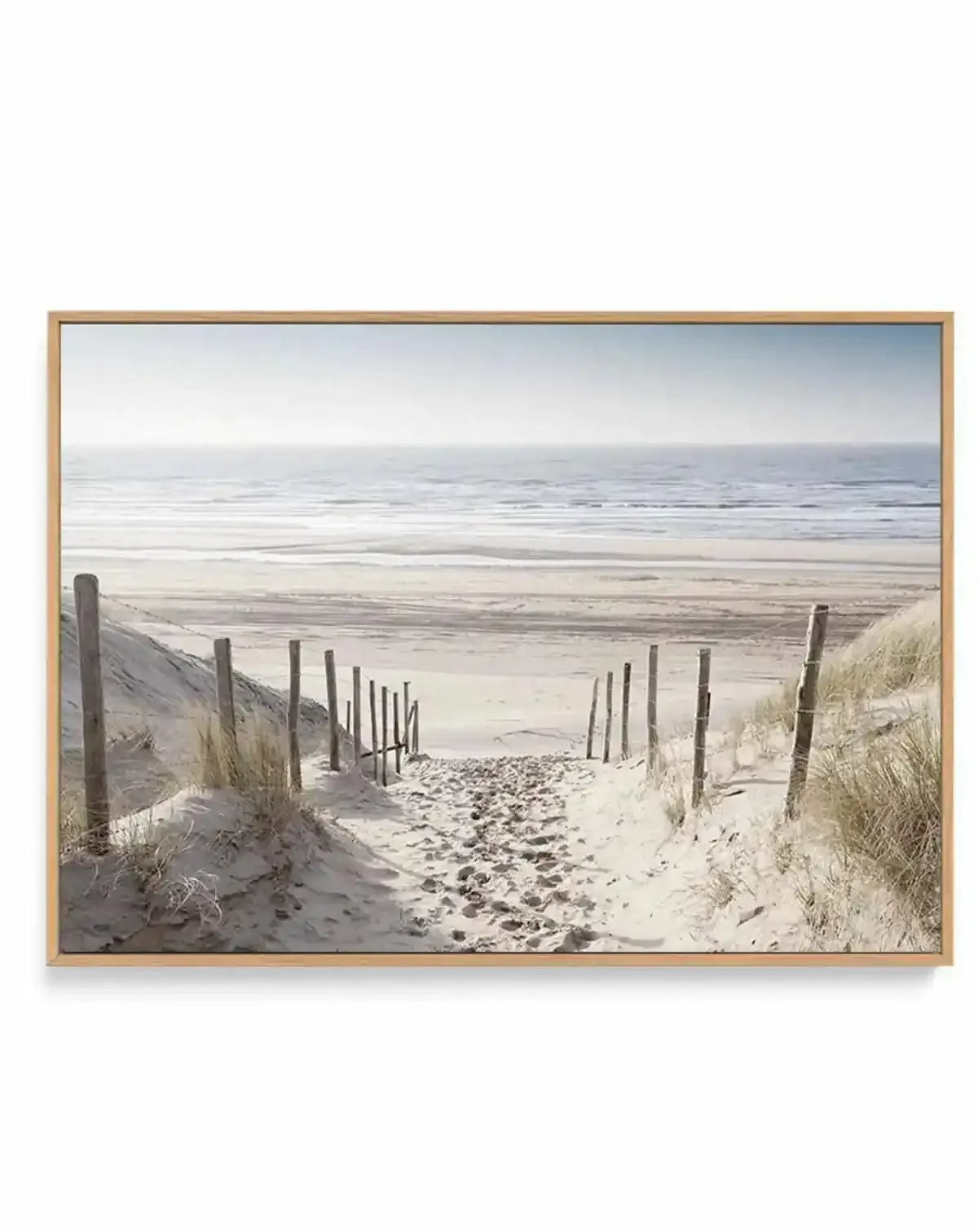 Framed canvas art print featuring a serene beach pathway scene. Wooden fence posts line a sandy path bordered by pale golden dune grasses on either side, leading toward a calm North Sea horizon. Soft, muted tones of cream, pale blue, and taupe dominate the composition. The photograph captures fine sand texture, weathered fence details, and the expansive flat coastal landscape under an overcast sky. The print is mounted in a natural wood frame with white border.